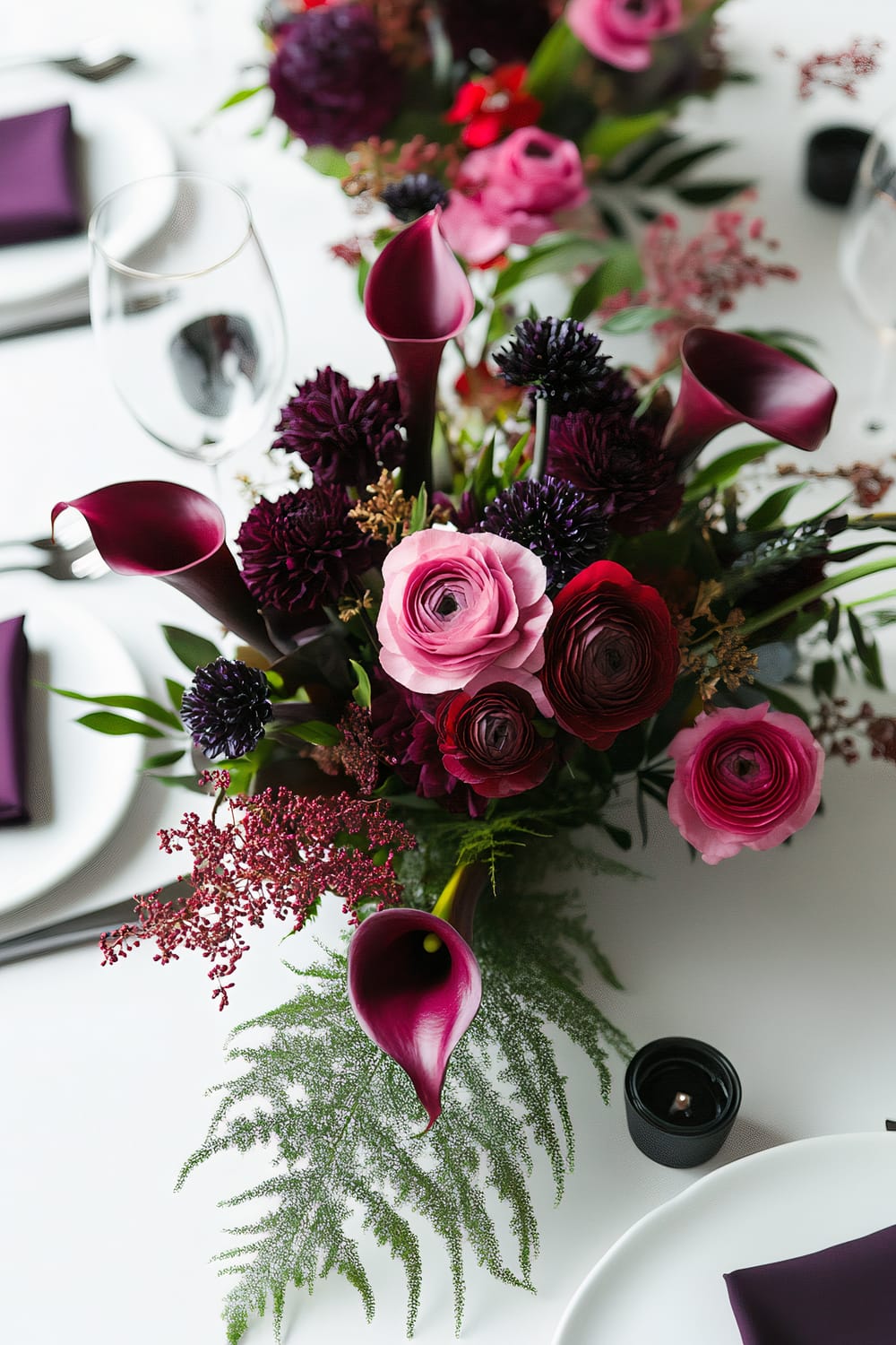 A table setting with a vibrant floral centerpiece featuring dark purple calla lilies, pink and magenta ranunculus, and deep burgundy blooms accented by fern leaves. A glass of water, white plates, and purple napkins are on the table.