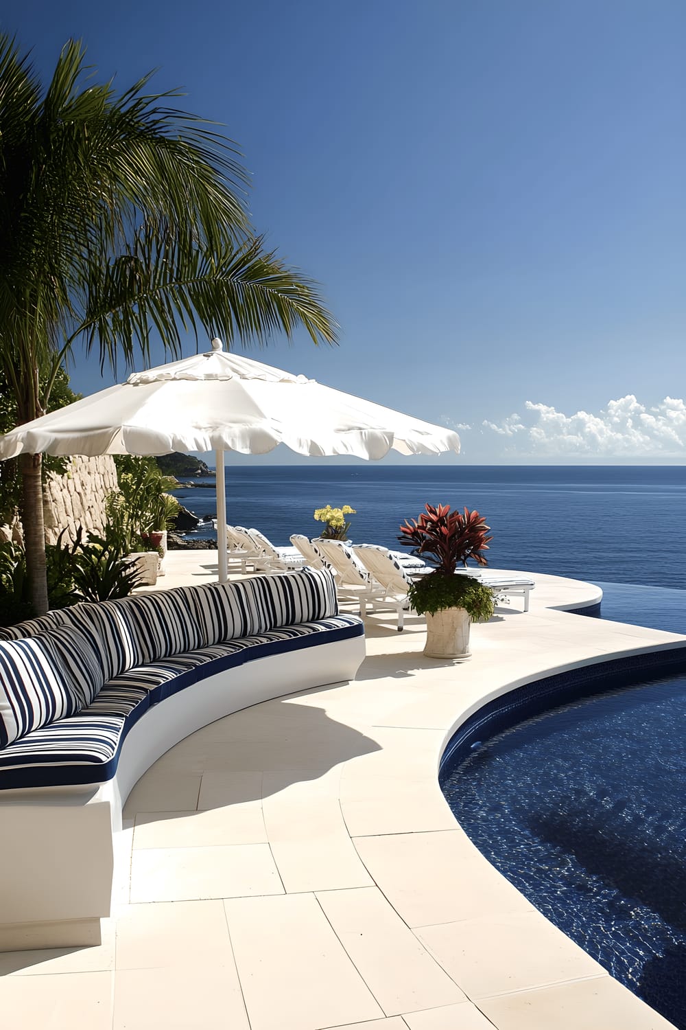 An image of a contemporary patio overlooking the ocean. The patio features white decking and an infinity edge pool. The furniture includes a curved outdoor sofa with navy and white striped cushions and a sun umbrella with scalloped edges providing shade. The patio is decorated with potted frangipani trees and tropical heliconias.