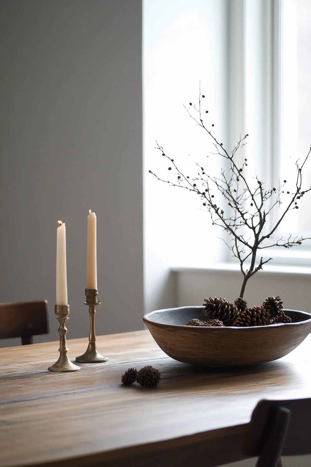 A minimalist wooden dining table with a rustic appeal, featuring two lit candles in brass holders and a large wooden bowl filled with pinecones and a bare branch. The setting is softly illuminated by natural light from a nearby window, creating a serene and understated ambiance.