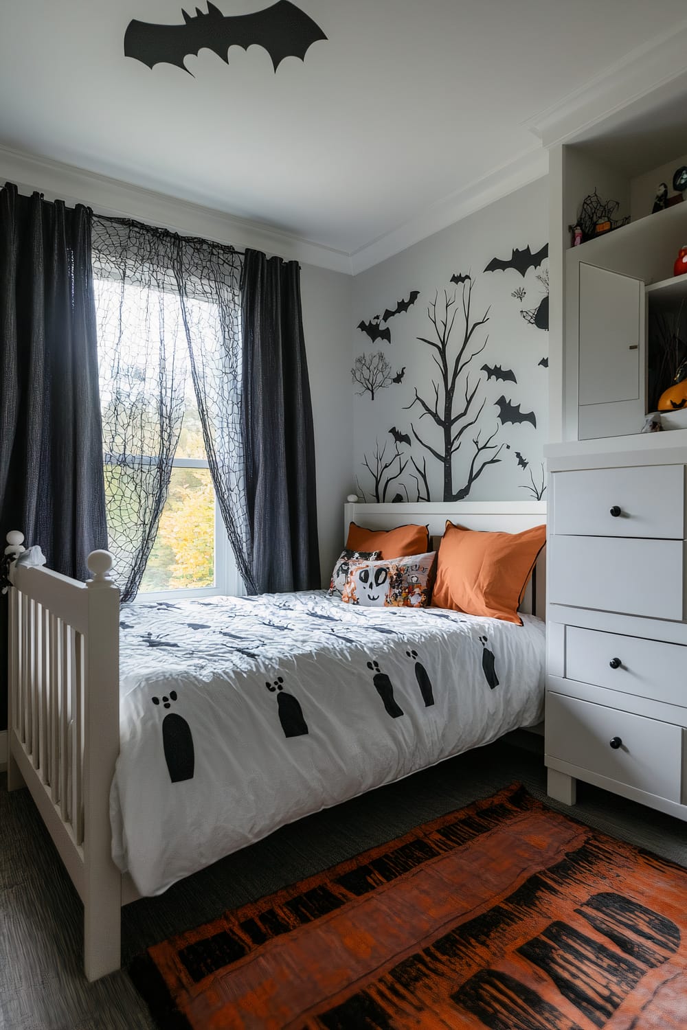 A small bedroom with a Halloween theme. The room has a white bed with orange pillows and a decorative pillow with a ghost design. The bedcover has black graveyard designs. The walls feature a tree and bats in black silhouette. A black spiderweb curtain hangs over the window. Above the bed, there is a large black bat decoration on the ceiling. A white dresser with black knobs stands next to the bed, and an orange and black rug lies on the floor.