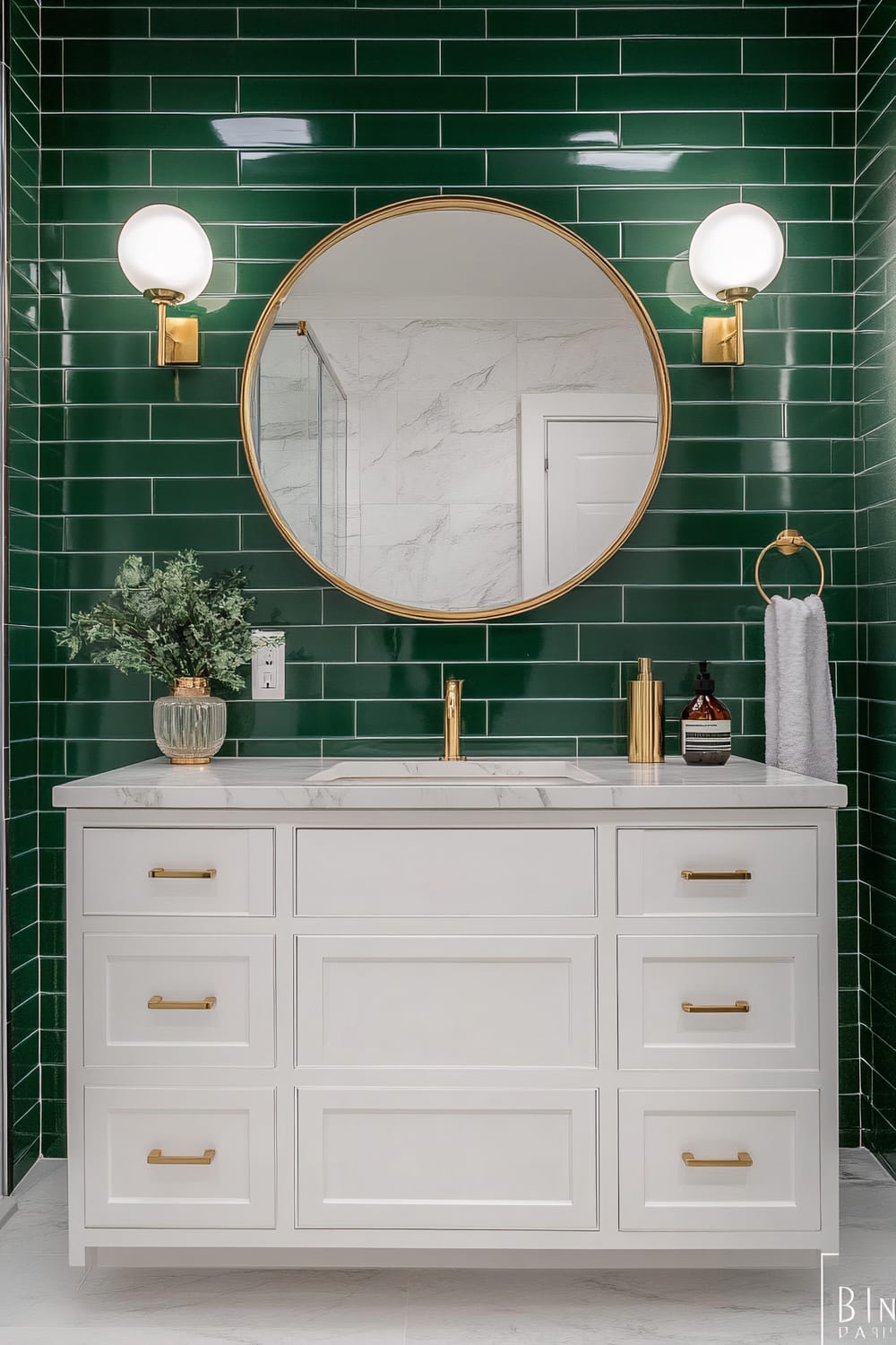 A bathroom features a modern vanity area with dark green tiled wall, white marble countertop, and brass fixtures. A round mirror with a brass frame hangs over the sink, flanked by two globe-shaped wall sconces. The vanity has white shaker-style drawers with brass handles. A potted plant in a glass vase, liquid soap dispenser, and folded towel complete the decor.