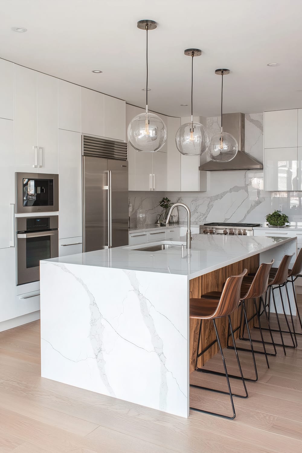 An elegant kitchen featuring white cabinetry with a glossy finish, integrated stainless steel appliances, and a large island. The island has a white marble waterfall countertop, a built-in sink, and offers wooden bar stools with metal legs. The backsplash and countertops also showcase the same marble. Above the island hang three clear glass pendant lights, adding a modern touch to the space. Light wood flooring ties the room together.