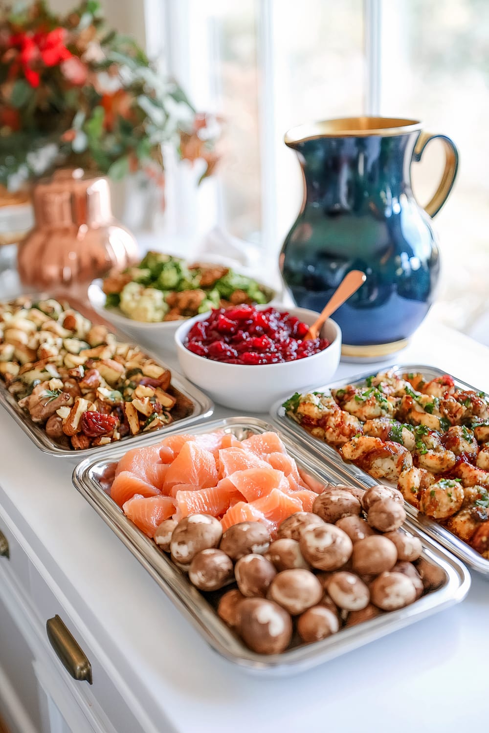 A Friendsgiving serving area with a minimalist white table that holds three metallic trays of assorted appetizers, including stuffed mushrooms, smoked salmon bites, and roasted nuts. A deep red bowl with cranberry dip is placed in the center. Decorative spoons are on each tray, and there is a cobalt blue ceramic pitcher filled with lemonade. There are autumnal decorations in the background.