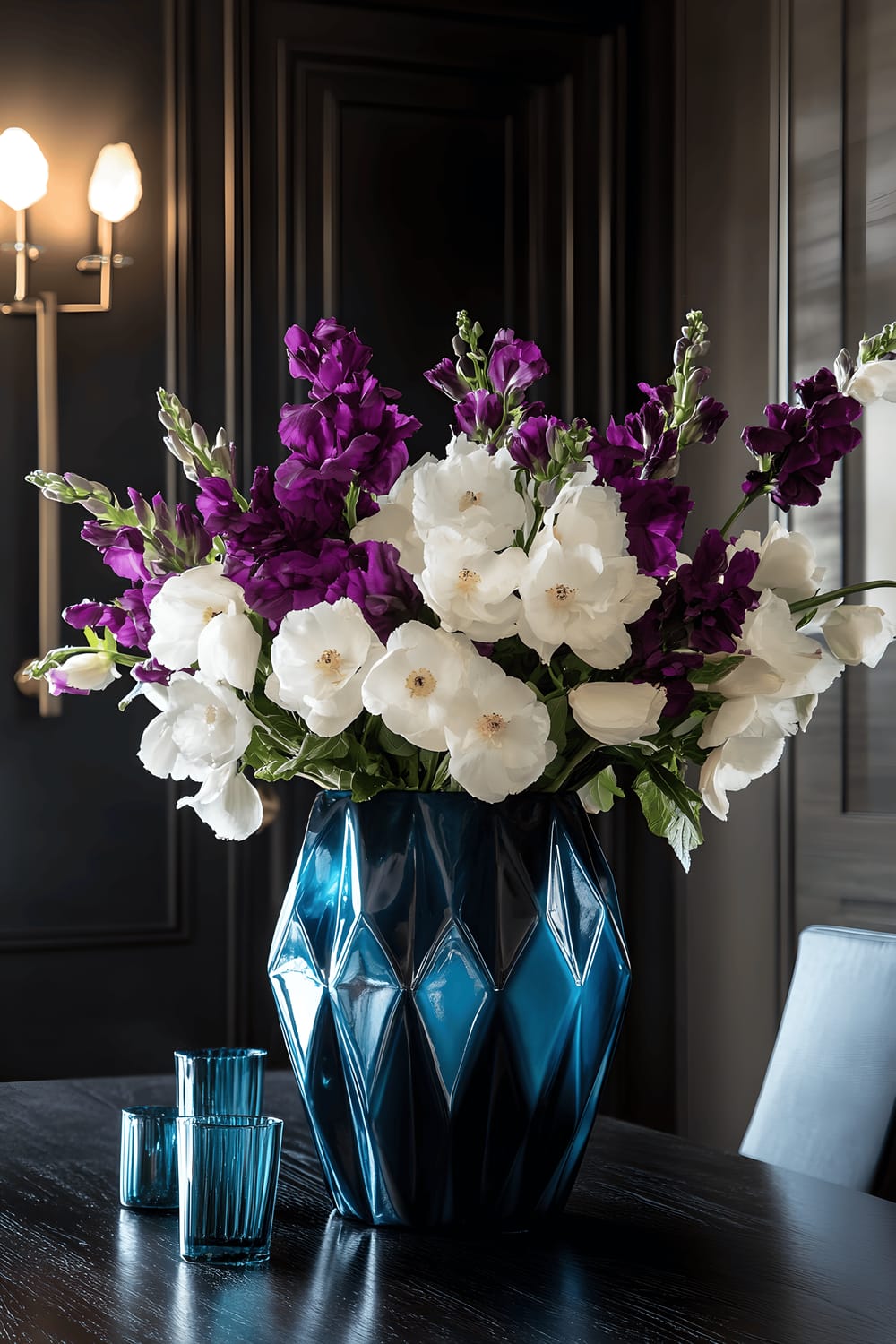 A minimalist dining room featuring a large cobalt blue geometric metal vase filled with white and purple flowers as table centerpiece. The vase is placed on a distressed wooden table with small pewter accents and teal glass ornaments nearby. The dining area is composed of dark walls and modern black chairs, highlighted by dramatic side lighting which accentuates the vividness of the vase's color and the sharpness of its angular design.