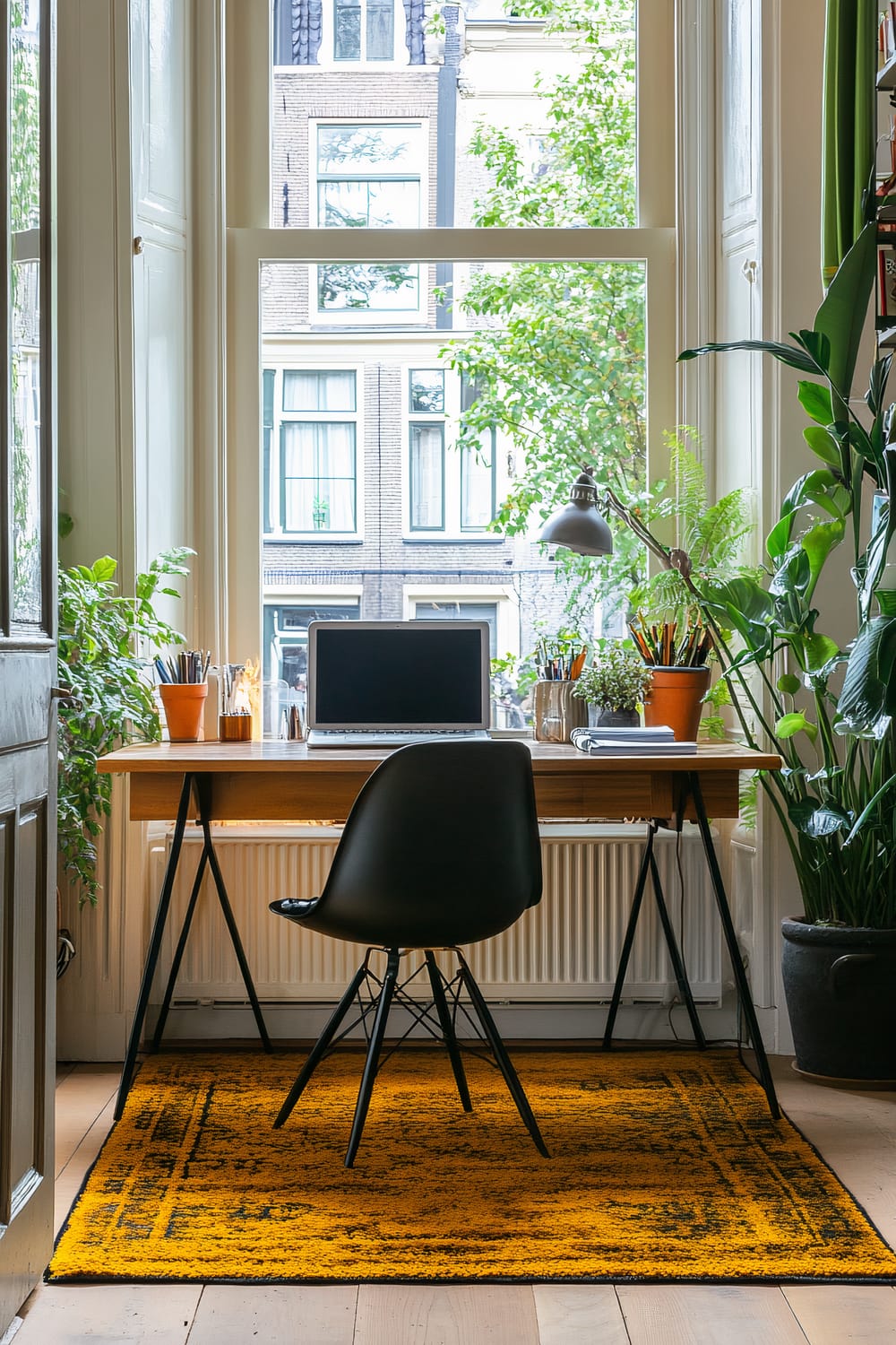 A home office setup in front of a large window. The desk has a wooden top and black metal legs, with a laptop, potted plants, and various writing utensils. A black chair is positioned in front of the desk, and there is a vibrant yellow rug underneath. Green leafy plants line both sides of the desk, and a large potted plant is on the floor to the right.