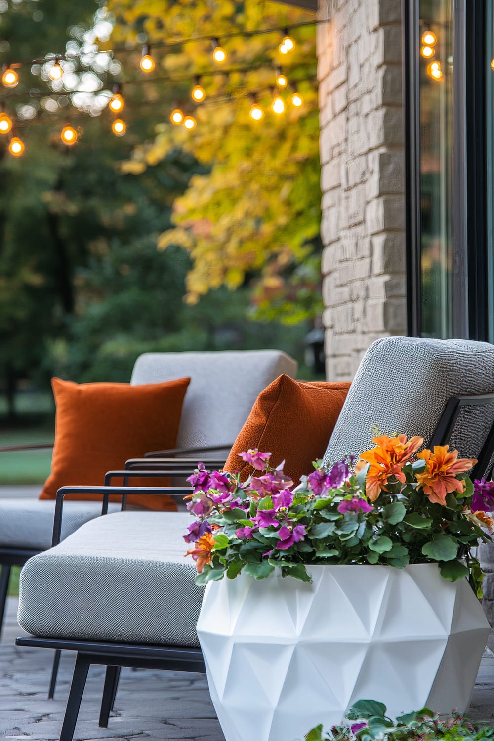 Outdoor seating area featuring modern gray cushioned chairs with burnt orange throw pillows. A geometric white planter brimming with colorful flowers sits beside the chairs. In the background, warm string lights hang above, and there are trees with autumn foliage visible.