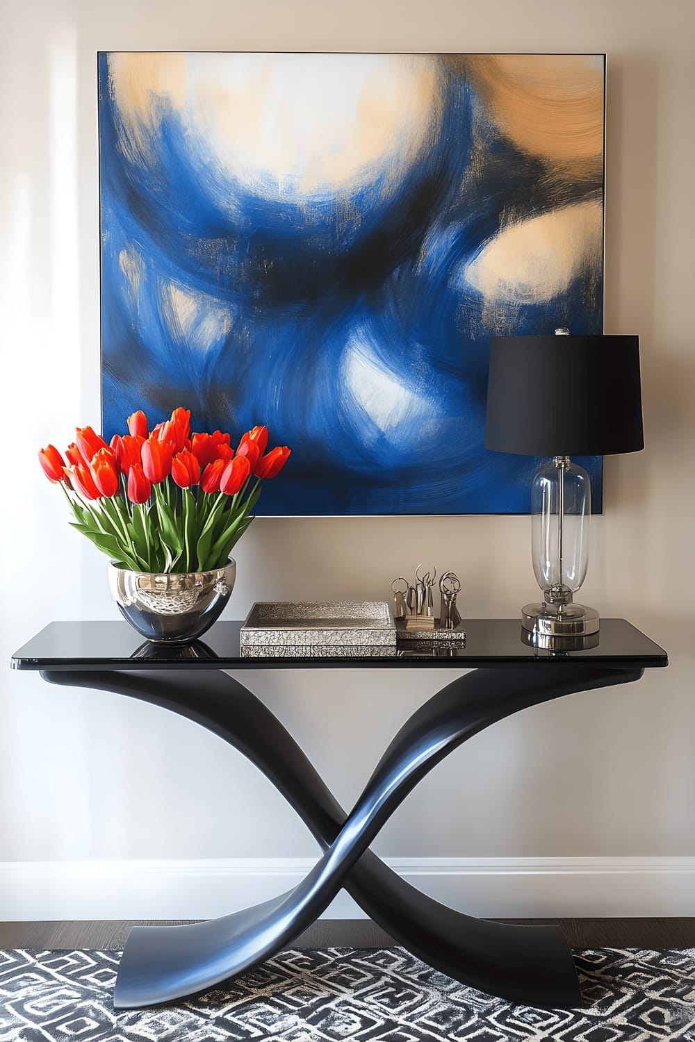 A stylish Milan entryway featuring a matte black metal sculptural console table with a glass top. The table is adorned with a decorative porcelain bowl filled with vibrant red tulips and a sleek silver tray holding keys and a modern lamp. Above the console is a large, abstract painting in shades of blue and gold. The floor is covered with a patterned black and white runner and there's a minimalist coat rack with leather hooks nearby.