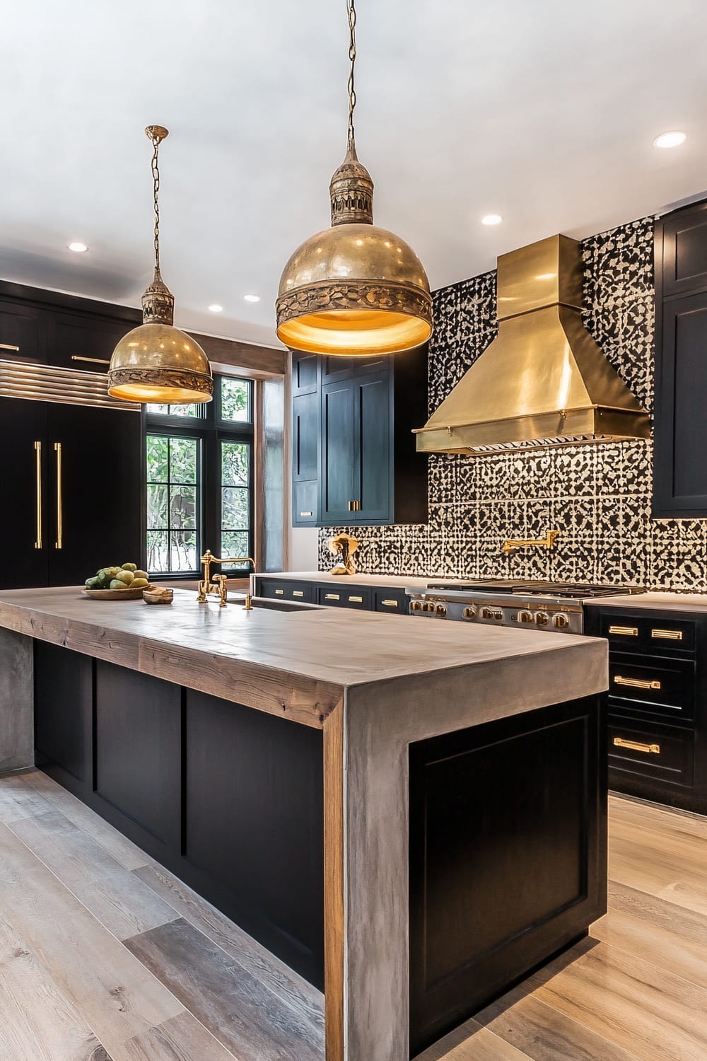 A kitchen featuring dark cabinetry, ornate brass pendant lights, and a large brass range hood. The backsplash showcases a black-and-white intricate tile pattern, and the island countertop is a mix of concrete and wood. Brass fixtures and hardware are present throughout, complementing the elegant and modern aesthetic.