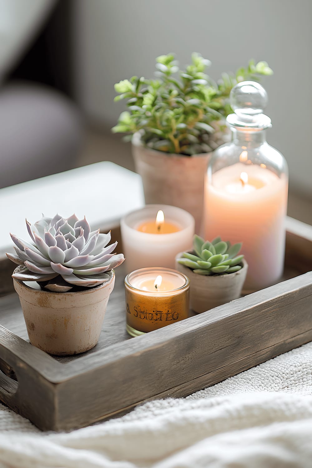 A reclaimed wooden tray displaying a collection of items for spring. The tray includes an arrangement of pastel-colored candles of different sizes, small potted succulents with green leaves, and an antique glass perfume bottle used as a tiny flower vase, holding some fresh pink blossoms.