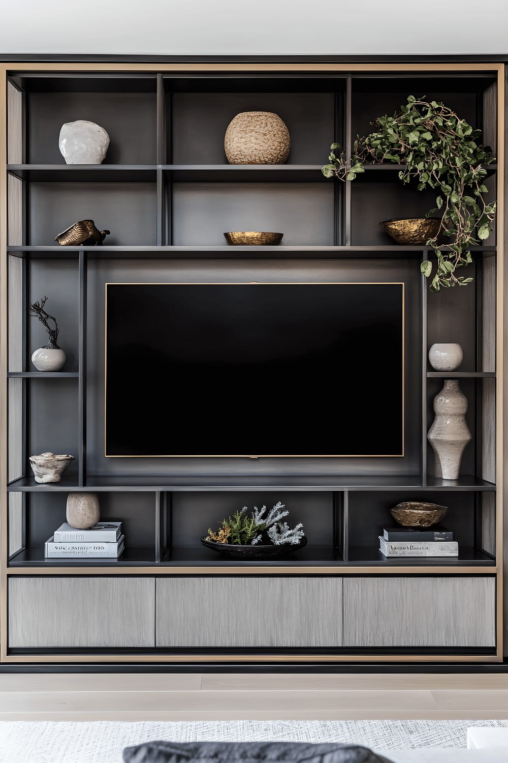 A large television is enclosed within a geometric metal frame with a brushed brass finish. It is flanked by minimalist black metal shelves adorned with carefully selected decorative pieces. The setup is illuminated by soft, natural light.