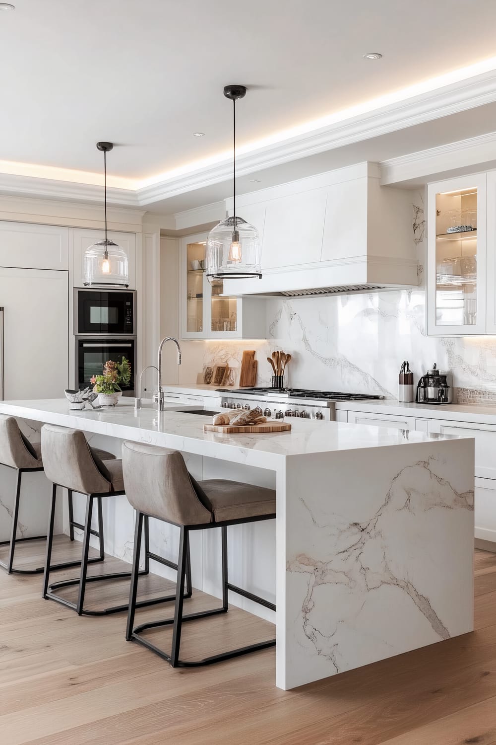 A bright, airy kitchen characterized by its modern design. The centerpiece is a large, marble-topped island with three gray cushioned bar stools. Overhead, two pendant lights with glass shades hang from the ceiling, emitting a warm glow. The kitchen features sleek, white cabinetry, including a built-in oven and microwave set, and a large, elegant marble backsplash. Wooden elements and stainless steel appliances add functional beauty. The soft, light-toned wooden floor complements the overall sophisticated aesthetic.