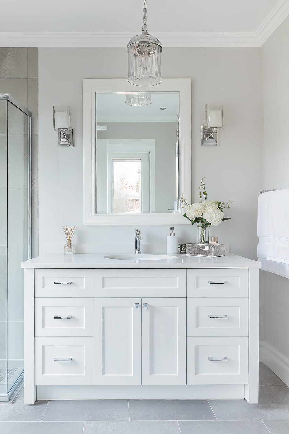 A modern bathroom featuring a white vanity with a seamless countertop and an undermount sink. Above the vanity hangs a large rectangular mirror with a white frame. Above the mirror, a glass pendant light fixture with metal accents is suspended from the ceiling. The vanity has multiple drawers and cabinets with sleek chrome handles. The walls are painted in a light gray color, complemented by matching gray floor tiles. A glass vase with white flowers and a silver tray with toiletries decorate the countertop. There is a glass-enclosed shower on the left side of the vanity.