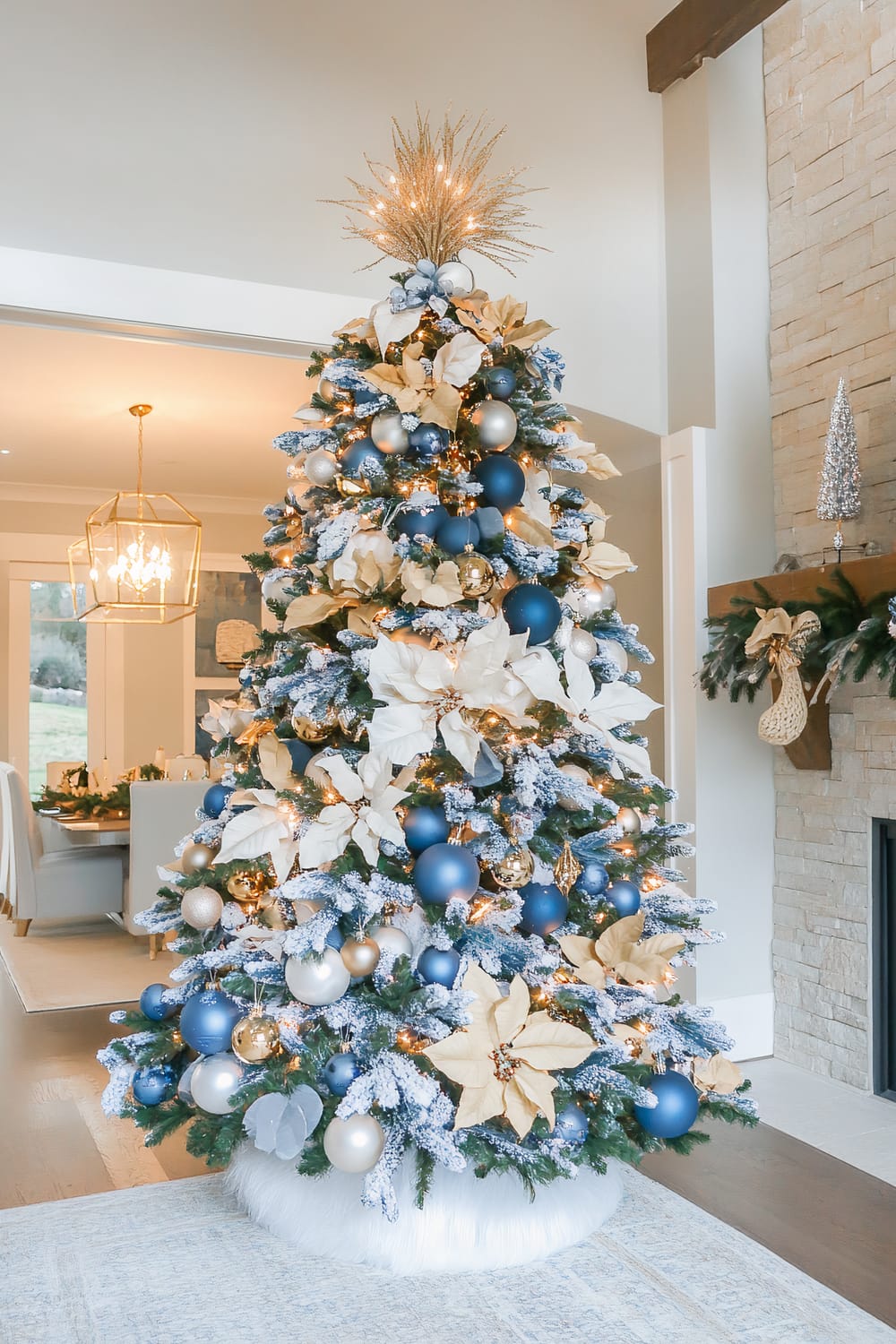 A living room decorated for the holiday season, featuring a tall Christmas tree adorned with blue, gold, and white ornaments, large golden poinsettias, and frosted branches. The tree topper is a golden, spiky starburst. In the background, a dining area is visible with a modern chandelier and an elegant dinner setting. A stone fireplace with a wooden mantle and festive decorations is also present.