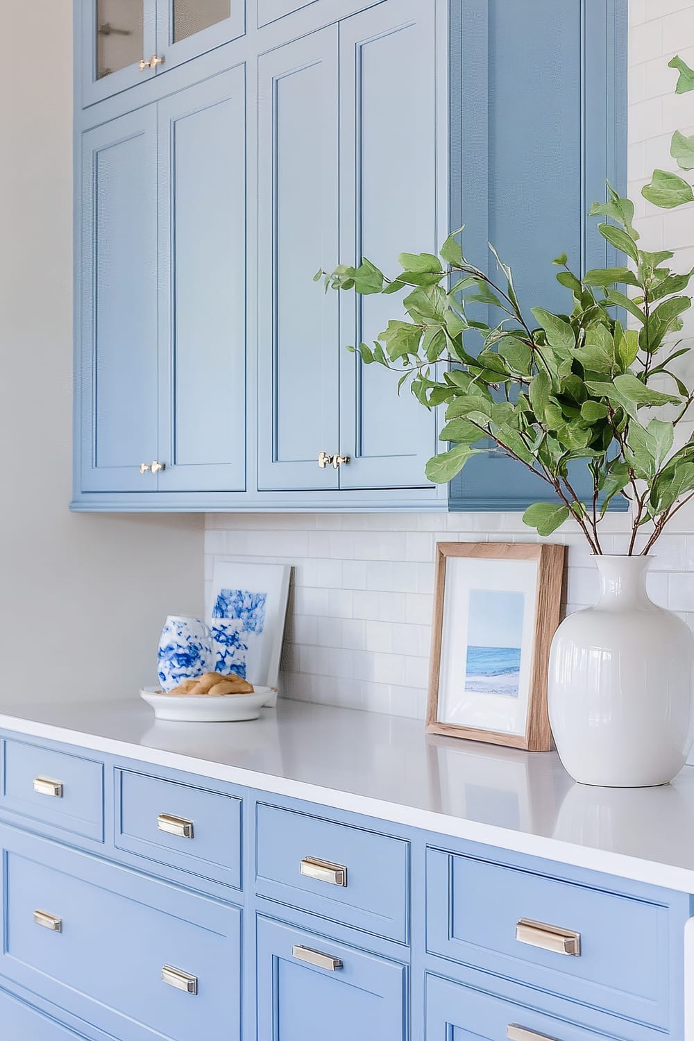 Interior shot showing light blue cabinets with brass handles and decorative items on the countertop, including a white vase with green foliage and framed photographs.