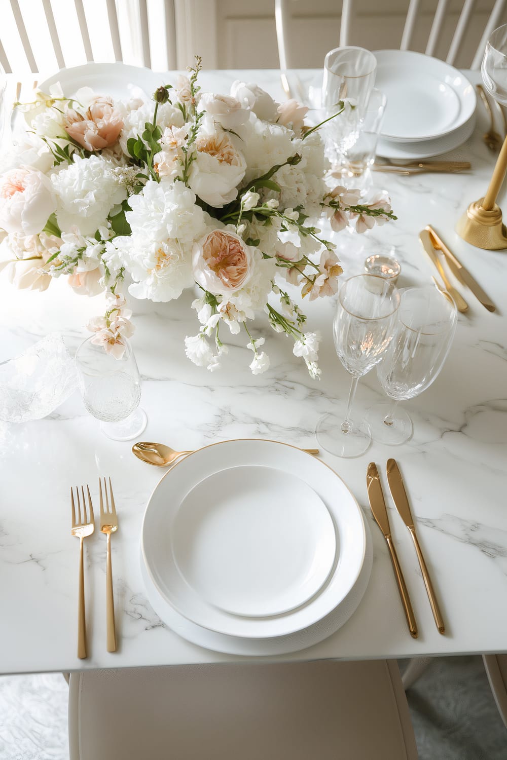 A sophisticated table setting featuring white porcelain plates stacked neatly atop a marble table. Gold-toned cutlery and crystal glassware, including water goblets and wine glasses, complement the pristine table setting. A lush floral centerpiece with cream, white, and pale pink flowers adds a touch of elegance.