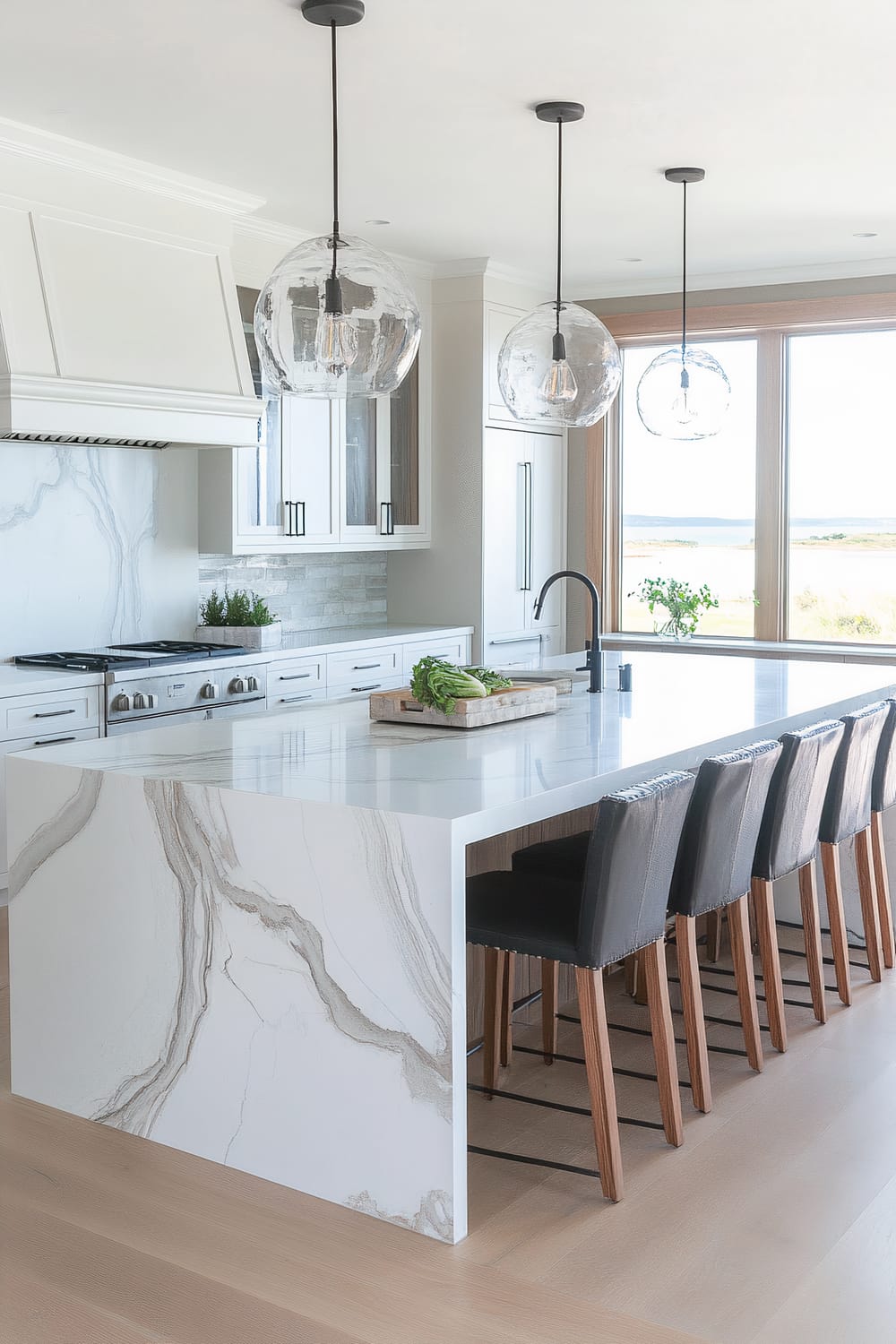 A contemporary kitchen with a large marble island featuring intricate grey veining. The island is paired with sleek bar stools with black upholstered seats and wooden legs. Three spherical glass pendant lights hang above the island. The kitchen is equipped with white cabinets, stainless steel appliances, and a black faucet. Large windows provide a scenic view of the outdoors.