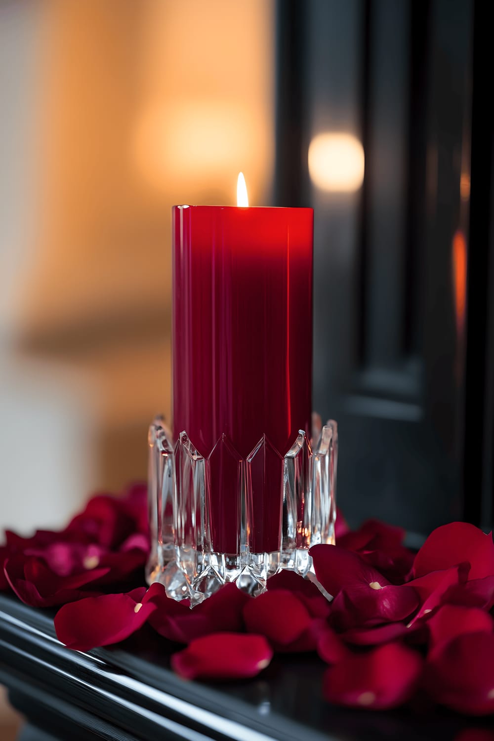 A deep red pillar candle, nestled in a clear crystal holder, forms the centerpiece on a black slate mantel. Fresh, scarlet rose petals are scattered around the candle, accentuating its glow. The softly lit surroundings evoke a romantic and elegant atmosphere.