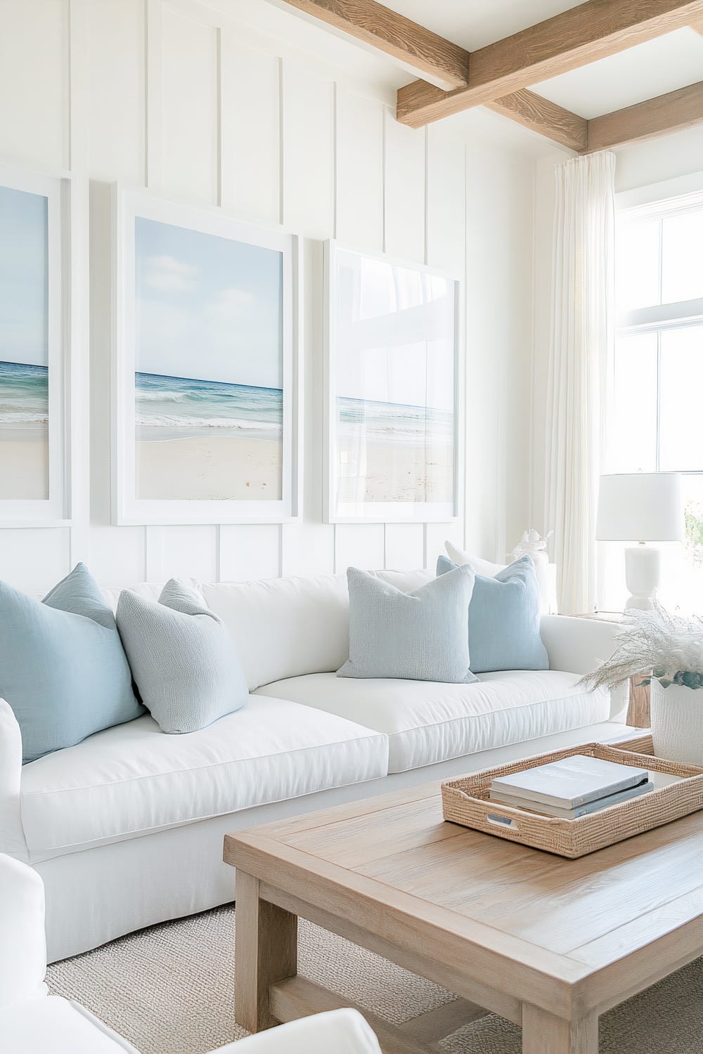 A bright and airy living room features a white couch adorned with light blue throw pillows. Above the couch hang three framed beach photographs, creating a serene coastal feel. A light wooden coffee table with a wicker tray containing books sits in front of the couch. The room has white, floor-to-ceiling wainscoting on the walls and large windows dressed with white curtains, letting in ample natural light. Decorative wooden ceiling beams add warmth and character to the space.