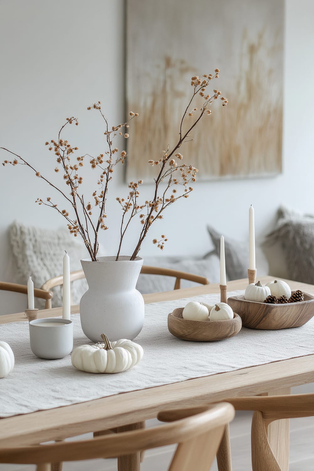 An elegant dining table setting features a minimalist approach with neutral tones. The table is adorned with a simple white table runner, a white vase with dried branches, wooden bowls containing white pumpkins and pine cones, and tall white candles in minimalist holders. A monochromatic painting with a nature theme hangs on the wall in the background, and cozy cushions are placed on the chairs.