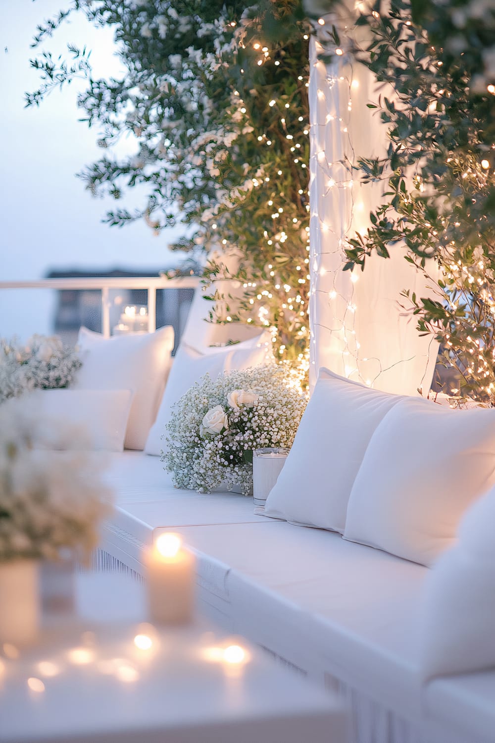 A serene white balcony decorated with enveloping white floral arrangements and softly glowing fairy lights. The seating area features plush white cushions and elegant holiday accents, enhancing the romantic atmosphere of the outdoor setting. The gentle candlelight adds to the cozy, tranquil evening ambiance.