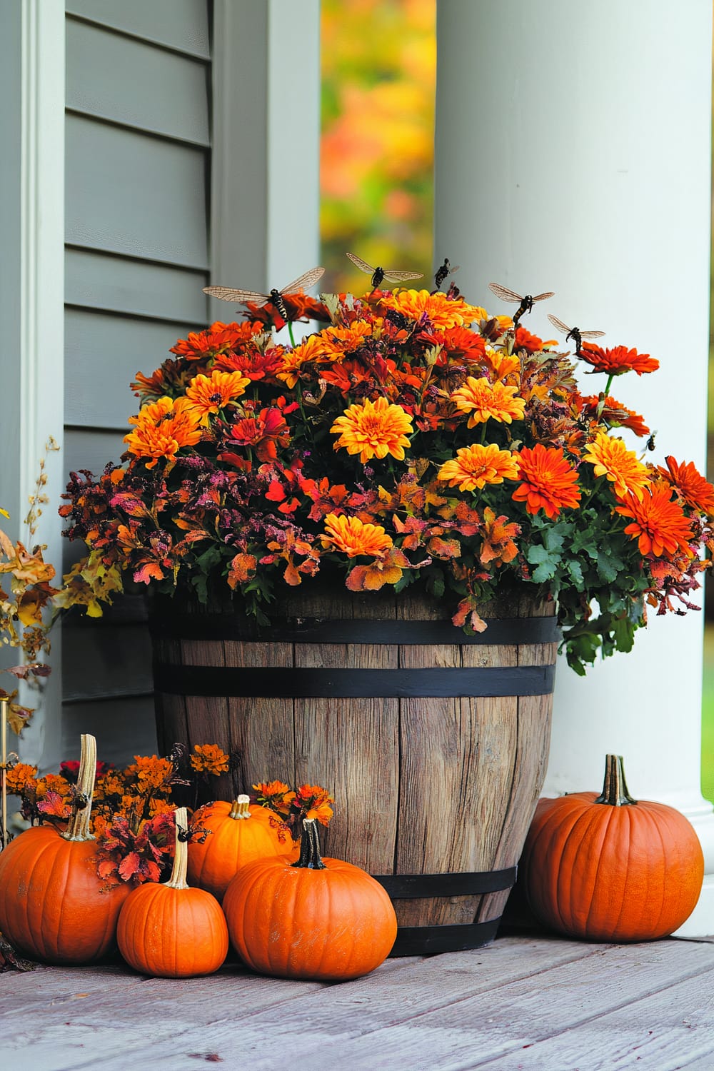 A rustic wooden barrel container filled with vibrant orange and yellow flowers stands on a wooden porch deck next to a vertical white column. Fall leaves and several pumpkins of various sizes are arranged around the base of the barrel. A few insects with transparent wings are hovering around the flowers and the pumpkins, adding to the natural and autumnal atmosphere. The background features the grey siding of a house and a softly blurred colorful autumn foliage.