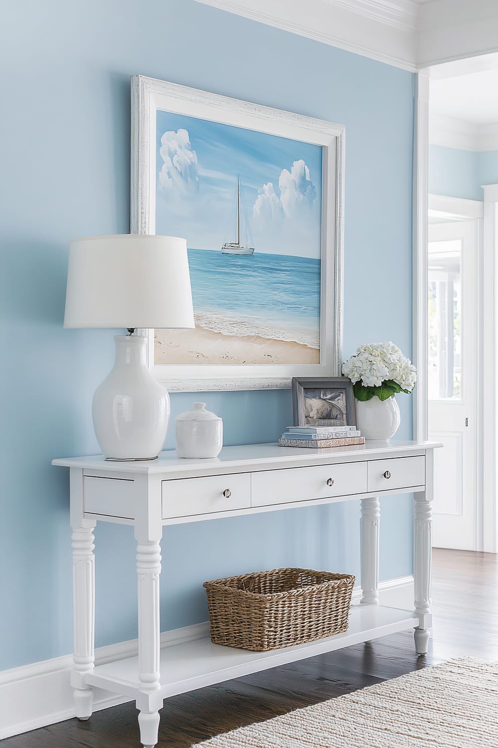 A coastal-themed entryway features a white wooden console table against a soft blue wall. The table is adorned with a white ceramic lamp and a few decorative items, including a small vase, books, a photo frame, and a vase with white hydrangeas. Above the table hangs a large painting of a sailboat on the ocean. The setup includes a woven basket on the lower shelf of the table, and the floor is dark wood with a light-colored woven rug.