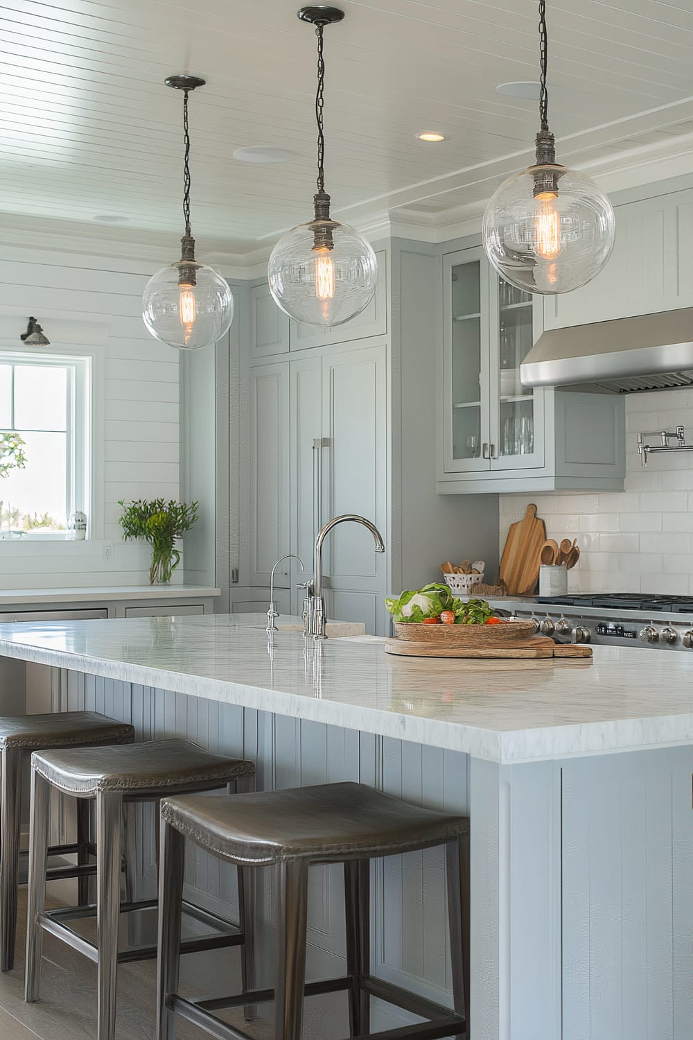 A modern kitchen featuring a sleek white marble countertop island with three gray bar stools underneath. Above the island hang three large globe pendant lights with visible filament bulbs. The kitchen has light gray cabinetry, stainless steel appliances, and a small window with a potted plant on the sill. A chrome faucet is installed on the island, and there is a wooden cutting board with vegetables on it.