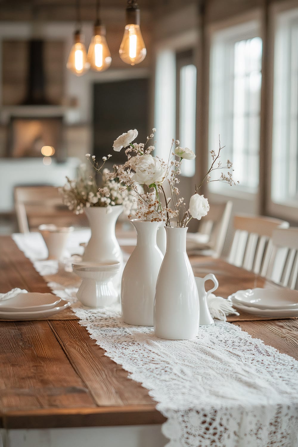 A rustic wooden farmhouse table is covered with a delicate white lace table runner. Several white ceramic vases, some holding white flowers, are arranged along the table's center. Warm pendant lights hang above, illuminating the table softly. The background includes wooden elements and large windows that allow natural light to flood the space.