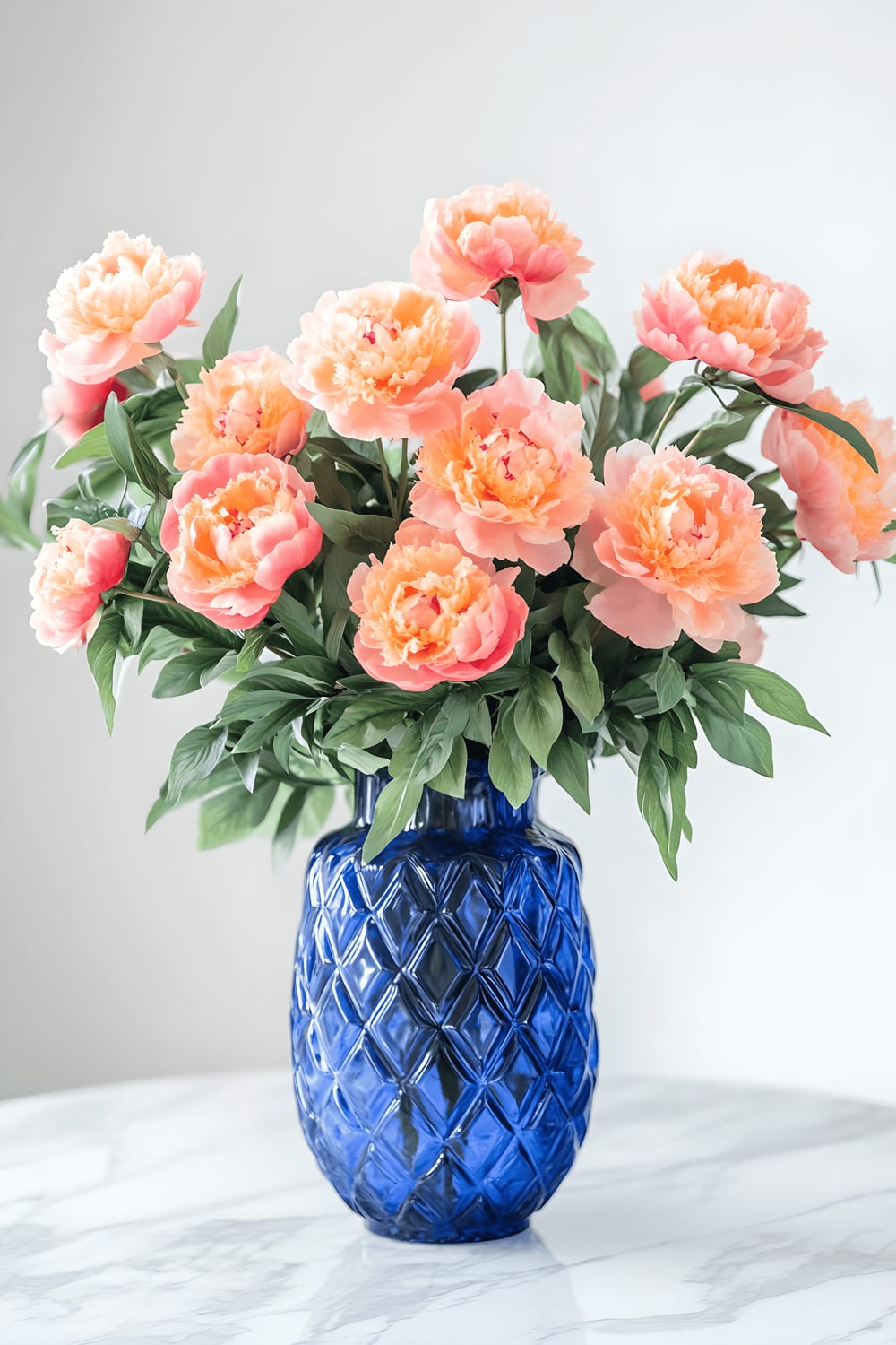 A large cobalt blue vase with intricate geometric patterns on a sleek white marble table serves as a striking centerpiece. The vase is filled with cascading peach peonies. The vibrant blue of the vase contrasts beautifully with the soft peach tones of the flowers. The surrounding space features minimalist Milanese decor, and the ambient natural light creates a dynamic interplay of colors and textures.