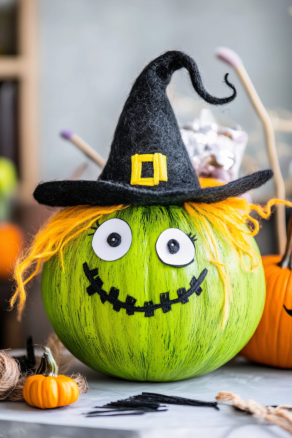 A green pumpkin decorated as a witch for Halloween. It has a black felt witch hat with a yellow buckle, orange yarn hair, large googly eyes, and a stitched smile. The background shows blurred objects, including a small orange pumpkin and some craft supplies on the surface.