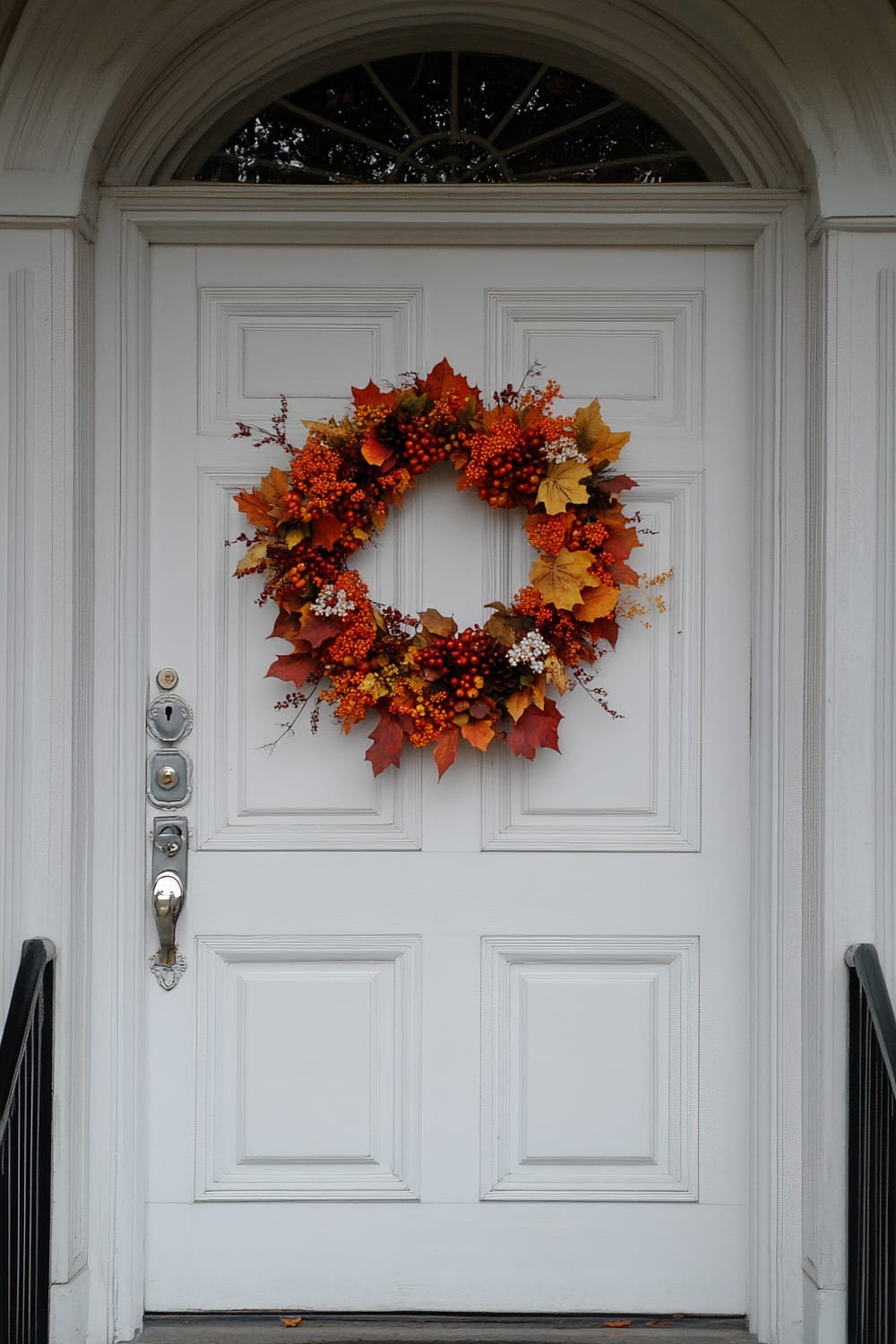 A white door adorned with an autumn wreath hangs in the center. The wreath is made of vibrant orange, yellow, and red leaves, along with orange berries and small white flowers. The door is framed by white wooden panels and features an arched window above. There is a metal door handle and lock on the left side of the door, with a small step and black metal railings leading up to it.