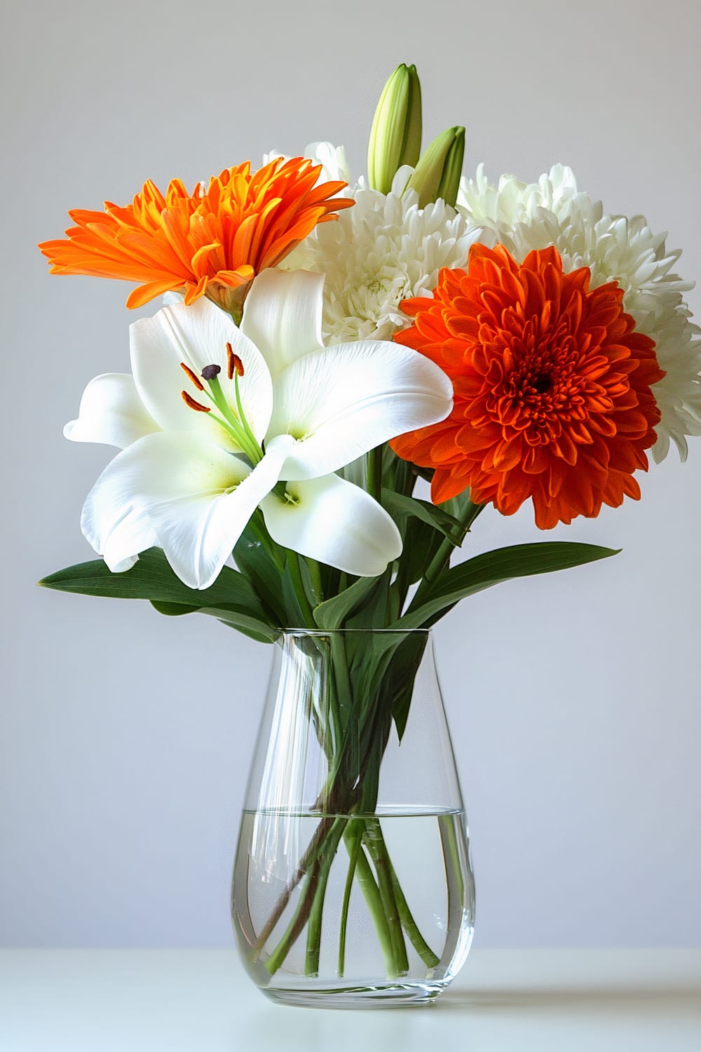 A clear glass vase containing a simple yet elegant floral arrangement is placed against a soft grey background. The bouquet consists of white lilies, orange gerbera daisies, white chrysanthemums, and green leaves, creating a vibrant and harmonious visual balance.