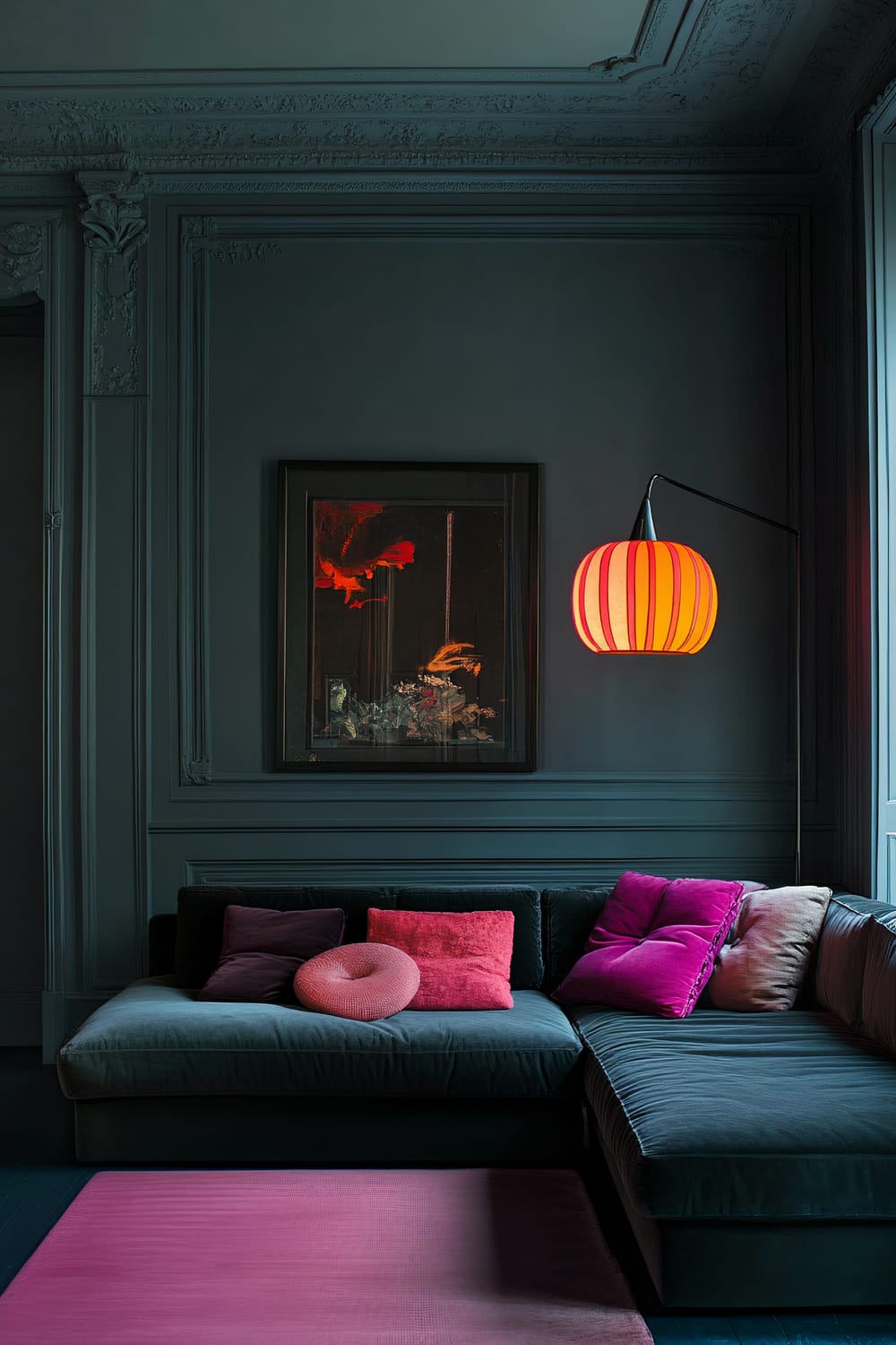 A French-inspired living room with walls painted in a blue-gray hue. The room features a dark velvet sectional sofa adorned with colorful cushions in shades of pink and red. A red and white striped lamp hangs on the wall above the sofa, casting a warm glow. A single statement artwork with red and black tones is displayed on the wall. Natural light streams in from a window, accentuating the vibrant colors in the space.