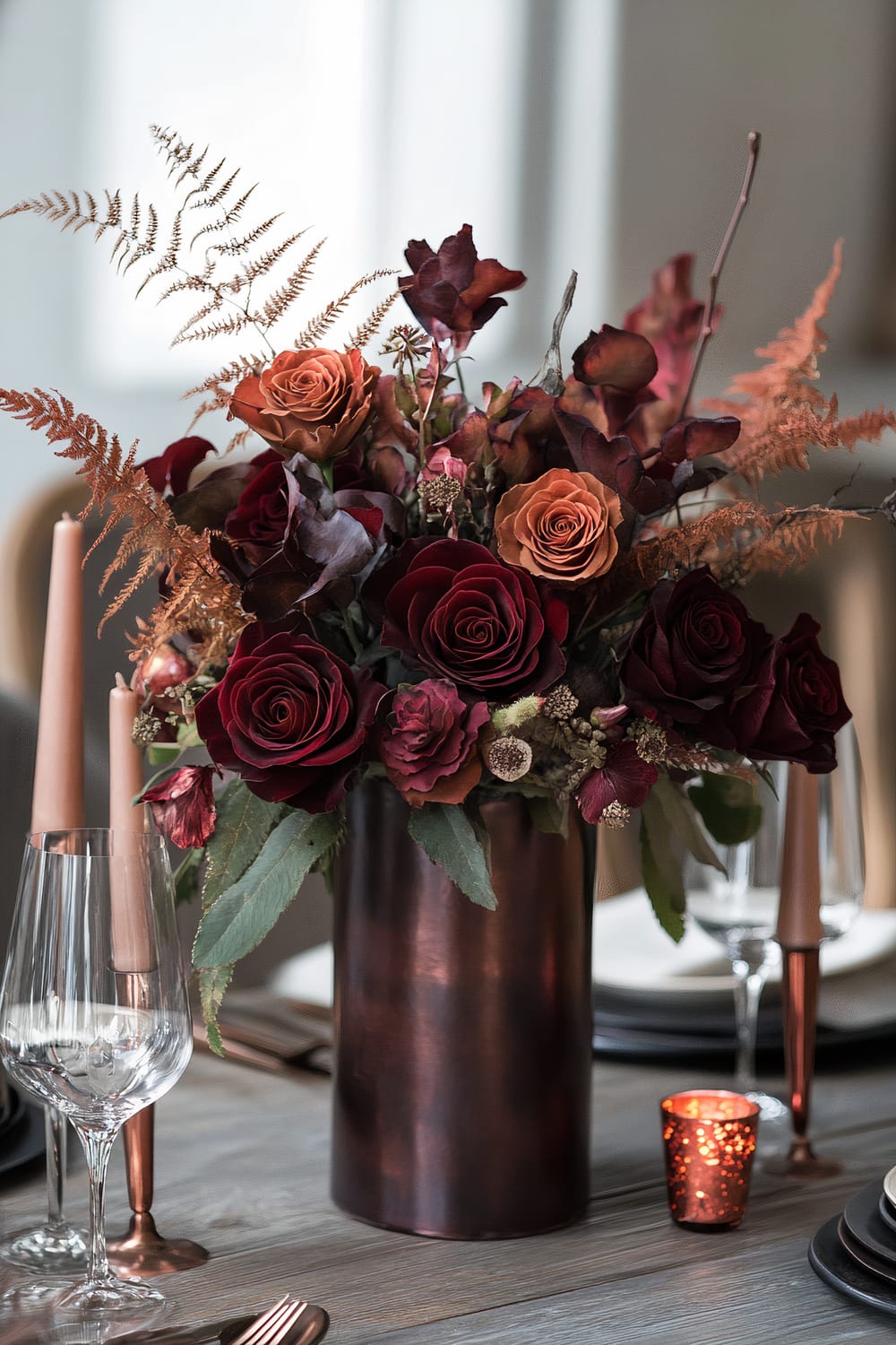 A table decorated with a large copper vase filled with a floral arrangement featuring deep red roses, orange roses, dried brown leaves, and ferns. The table also includes rose gold-colored candles in candleholders, glassware, and a small votive candle holder with a lit candle.