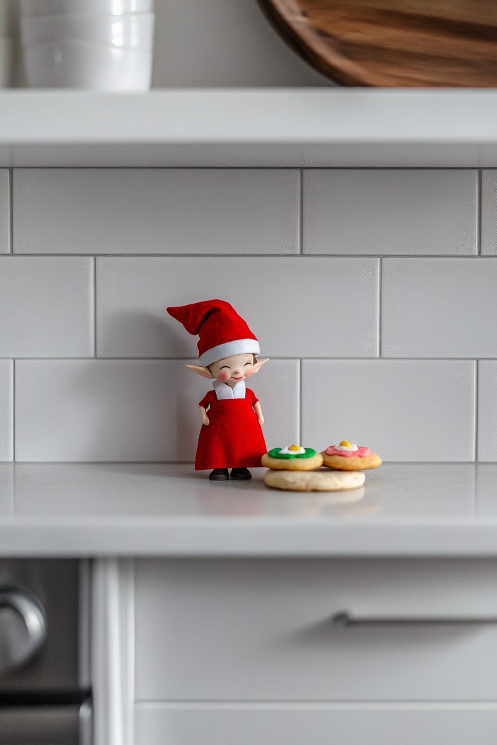 A small elf dressed in a red chef outfit stands on a white kitchen shelf. The elf is wearing a tiny red chef hat and apron, and appears to be placing two colorful cookies, one with green icing and the other with pink icing, on the shelf. The background consists of light gray tiled walls with soft natural lighting.