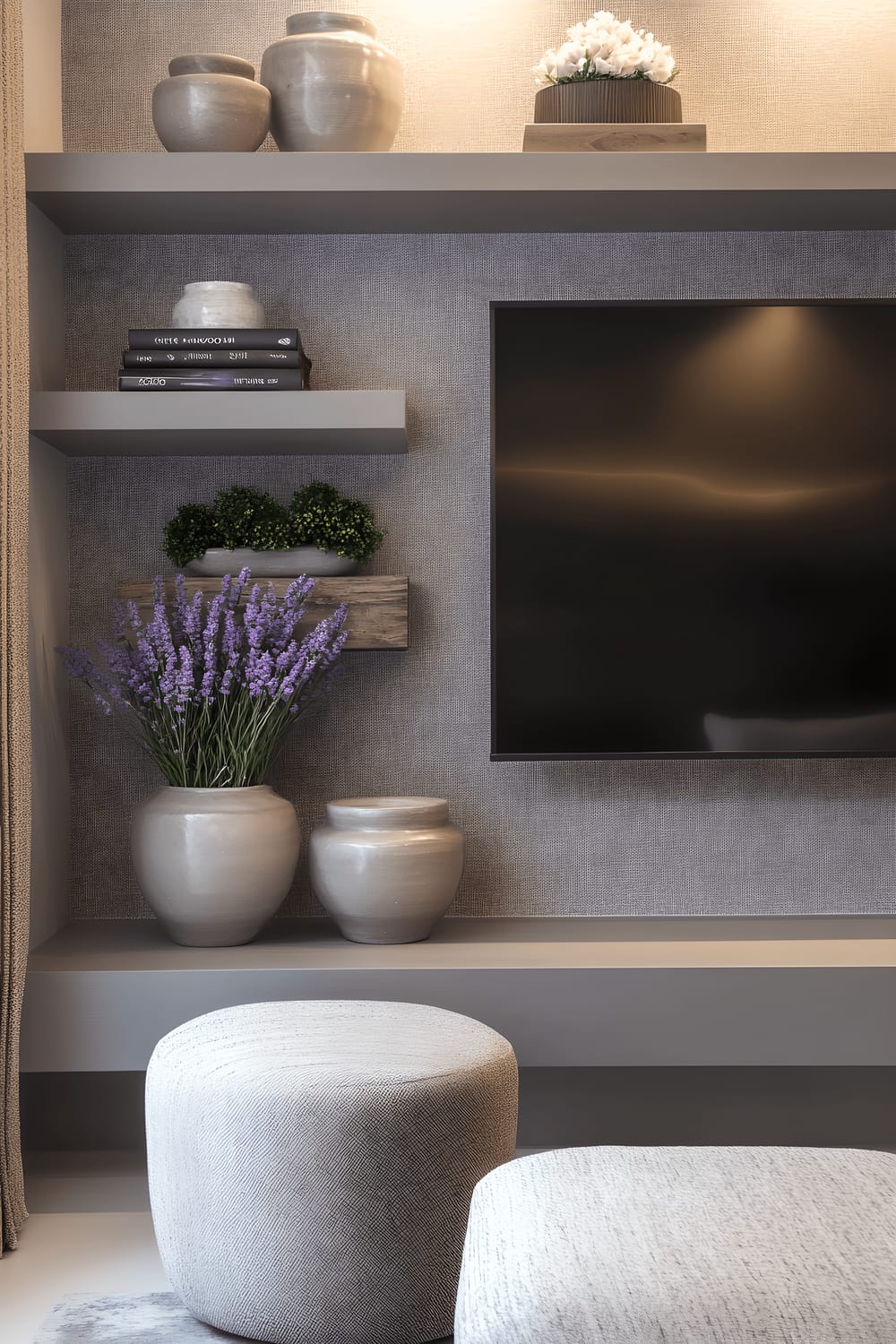 A television wall decorated in calm tones of lavender and grey. The wall displays a clean, modern TV set, surrounded by floating grey shelves boasting stylish decor items like lavender-scented candles and ceramic objects. The backdrop sports a soothing lavender and light-grey wallpaper, concluding with soft, relaxing illumination achieved through lavender-toned light fixtures.