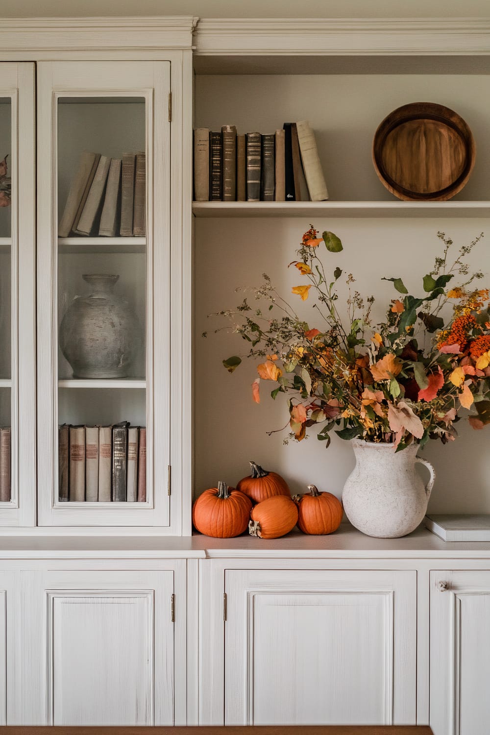 A white wooden cabinet with glass panes and an assortment of items on its shelves. The left side of the cabinet displays vintage books and a weathered vase behind glass doors. An open shelf on the right features neatly arranged old books, a round wooden tray, four small pumpkins, and a white speckled vase filled with an organic arrangement of autumn leaves and flowers.