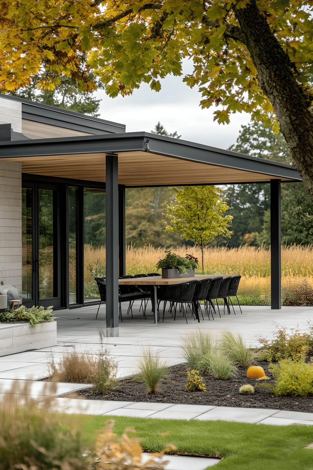 A modern outdoor patio featuring a wooden dining table with sleek black chairs, set underneath a vibrant spring tree. The gray tile flooring mirrors a clean aesthetic, along with lattice privacy walls. Minimalist planters filled with lush greenery contrast beautifully with the contemporary design, while a stainless steel grill rests to the side. Decorative pumpkins add a hint of seasonal charm to the space.