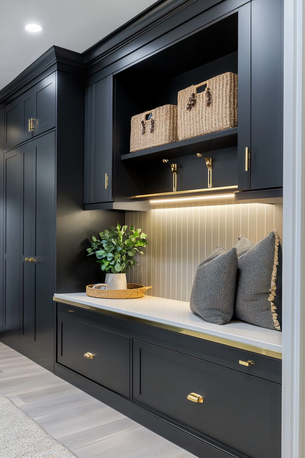 An elegant mudroom scene showcasing built-in black cabinetry with gold hardware. There are wicker baskets on an open shelf, a built-in bench with throw pillows, and a potted plant on the bench. LED strip lighting illuminates the space underneath the upper cabinets. The flooring is a light wood, complementing the dark cabinetry and creating a balanced contrast.