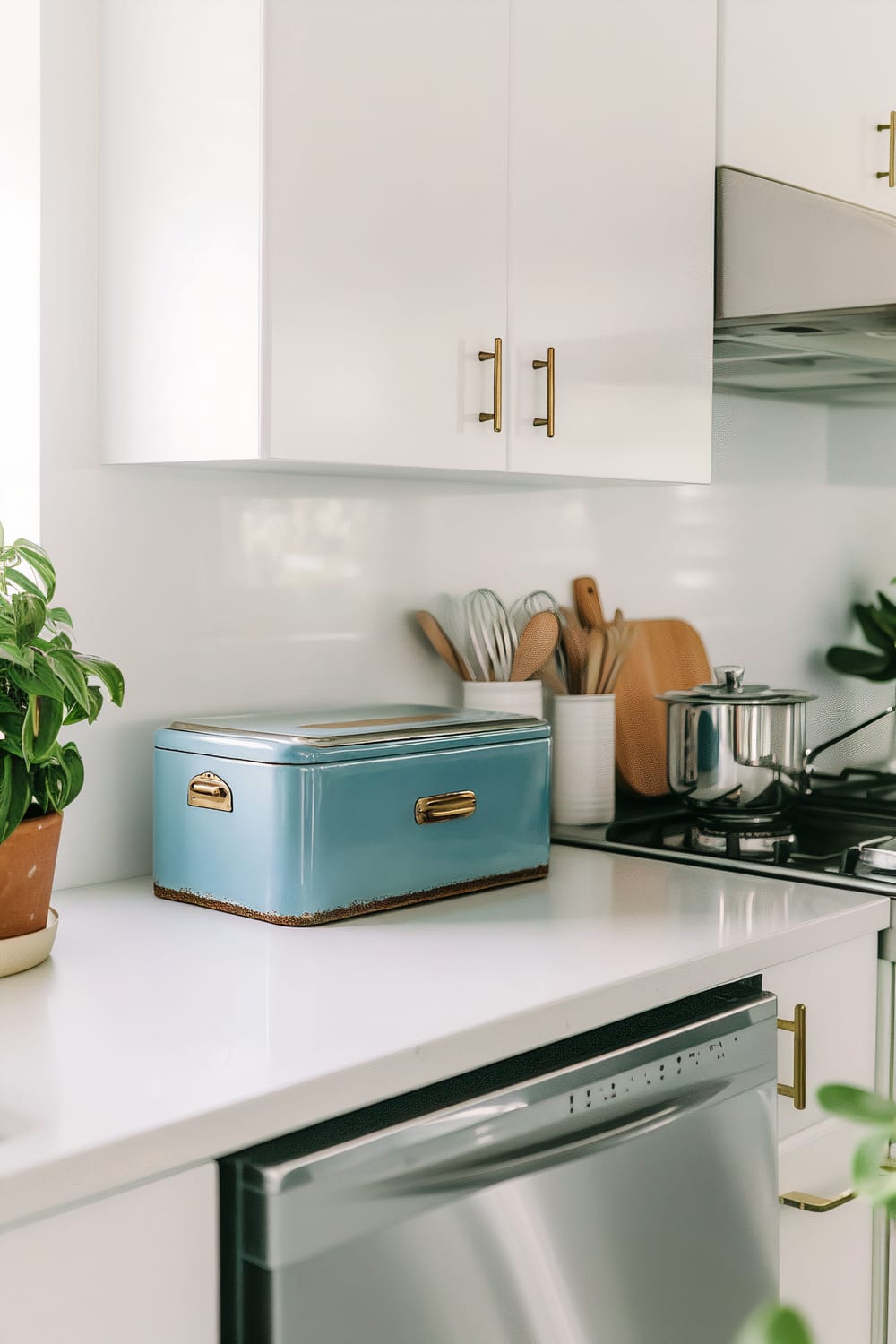 An image of a contemporary kitchen countertop with a sleek, white quartz surface. On the countertop, there is a vintage-style blue metal bread box with brass handles showing slight rust for a vintage touch. To the right, wooden kitchen utensils are neatly stored in a white ceramic holder, next to a stainless steel pot on a gas stove. There is a potted plant with lush green leaves to the left of the bread box, bringing some natural elements into the space. The white, minimalist upper cabinets have brass handles, reflecting the ambient light softly.