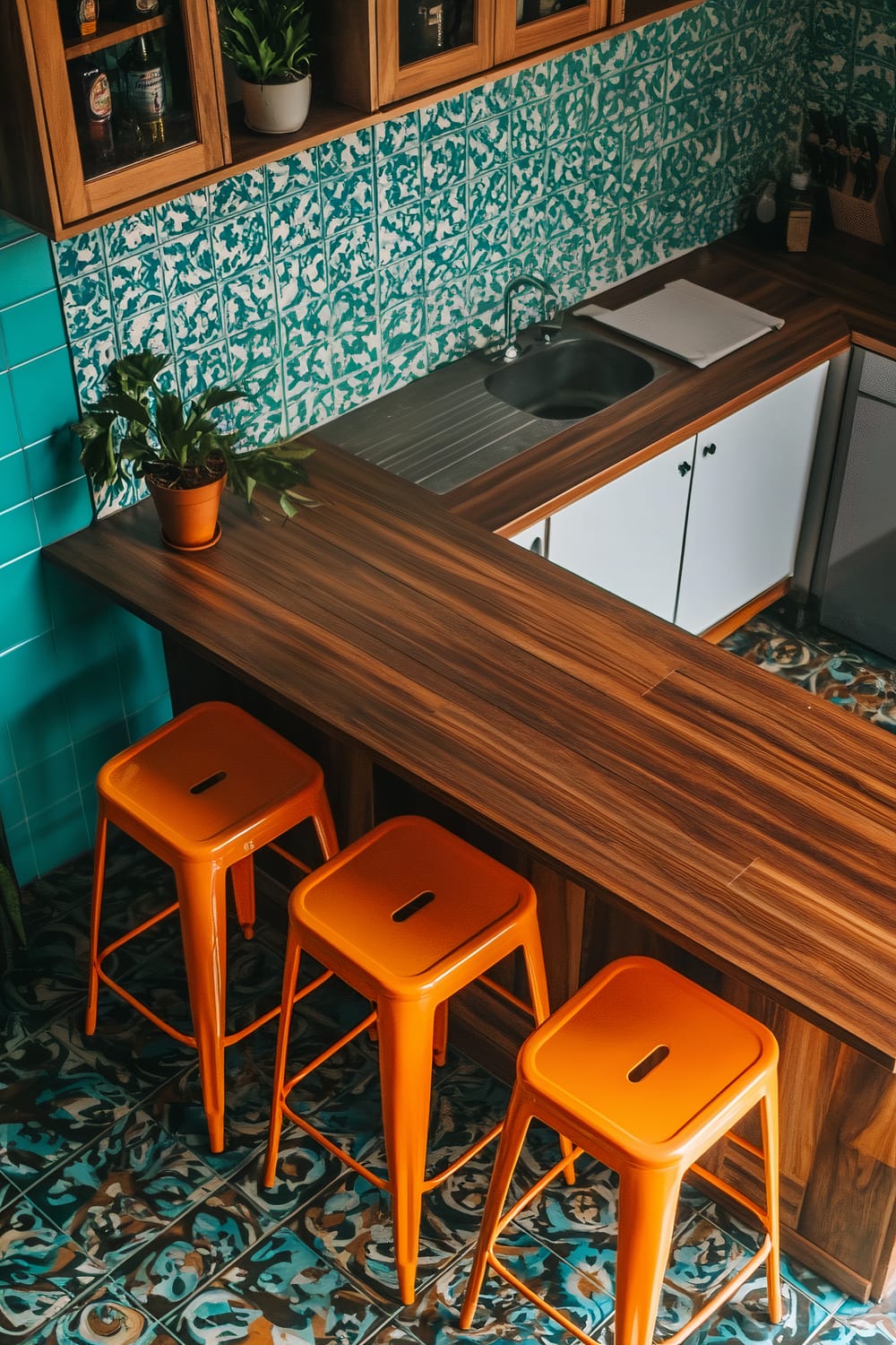 An overhead view of a 1970s kitchen features a brown wooden countertop kitchen island with three orange plastic bar stools. The teal patterned backsplash complements the flooring, and the space includes a small sink, potted plants, and warm wooden cabinetry.