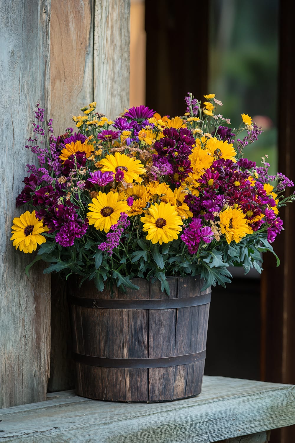 A wooden bucket filled to the brim with vibrant, freshly picked flowers is placed on a rustic ledge. The arrangement includes a mix of yellow, purple, and dark red flowers, creating a colorful and lively display. The backdrop consists of weathered wooden planks and a partial view of a door, giving the scene a cozy, country feel.