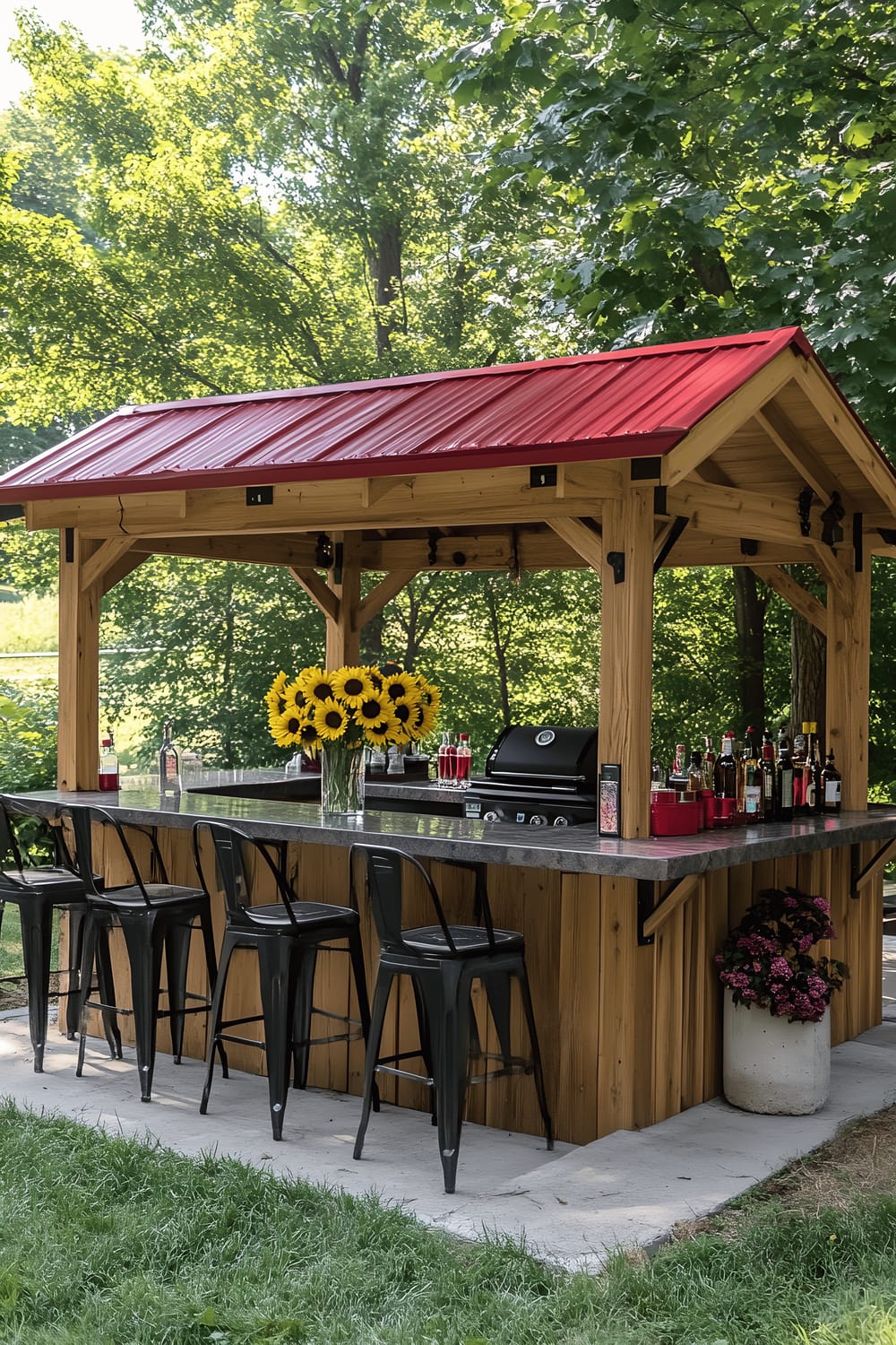 A spacious backyard featuring a wooden gazebo with a red metal roof housing an inbuilt bar and grill area. There is seating around the counters with black bar stools. On the counter, there's a vase with sunflowers alongside casual dining setup. The gazebo stands on a concrete base against a backdrop of lush greenery.