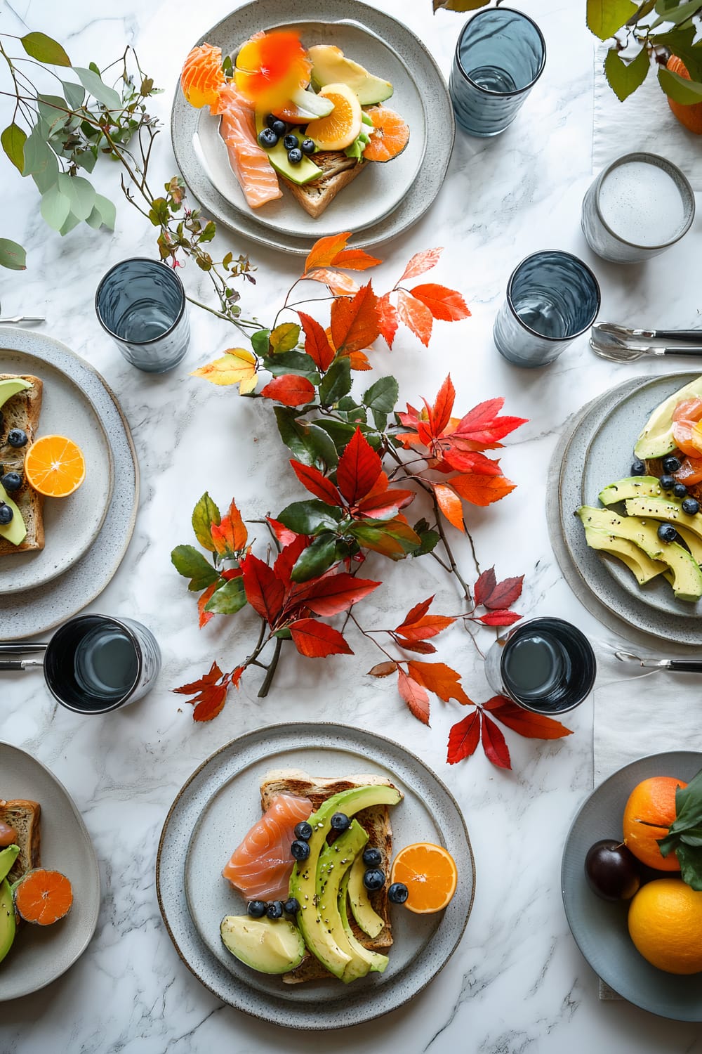 A minimalist Friendsgiving brunch table set on a white marble surface. Six earth-toned ceramic plates each hold avocado toast, smoked salmon, and fresh fruit salad. A geometric metallic centerpiece in deep yellow with red autumn leaves and small branches is placed in the center, accompanied by a silver metallic vase filled with fresh greenery. Five vintage-inspired cobalt blue glass mugs filled with herbal tea are evenly distributed around the table. Striking pendant lighting illuminates the scene against a white backdrop adorned with subtle autumn foliage.