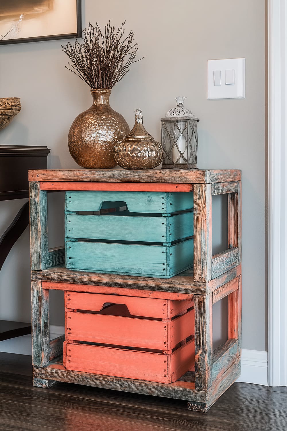 A stack of repurposed wooden crates, painted in bright coral and turquoise, is used for storage in a welcoming entryway. The crates are against a light grey wall and illuminated by dramatic lighting. Decorative items like vases and a lantern are placed on top of the crates.