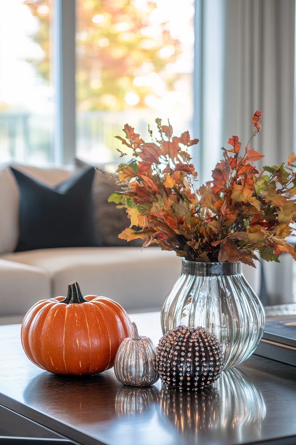 A sophisticated autumn-themed display is positioned on a dark wooden coffee table in a modern living room. The centerpiece features a clear ribbed glass vase containing an arrangement of colorful autumn leaves in hues of orange, red, and green. Surrounding the vase are three decorative pumpkins: one is a standard orange pumpkin, the second is a shiny silver mini-pumpkin, and the third is a black pumpkin encrusted with silver rhinestones. In the background, a cream-colored sofa with black and brown throw pillows is visible, along with large windows showcasing the blurred colors of fall foliage outside.