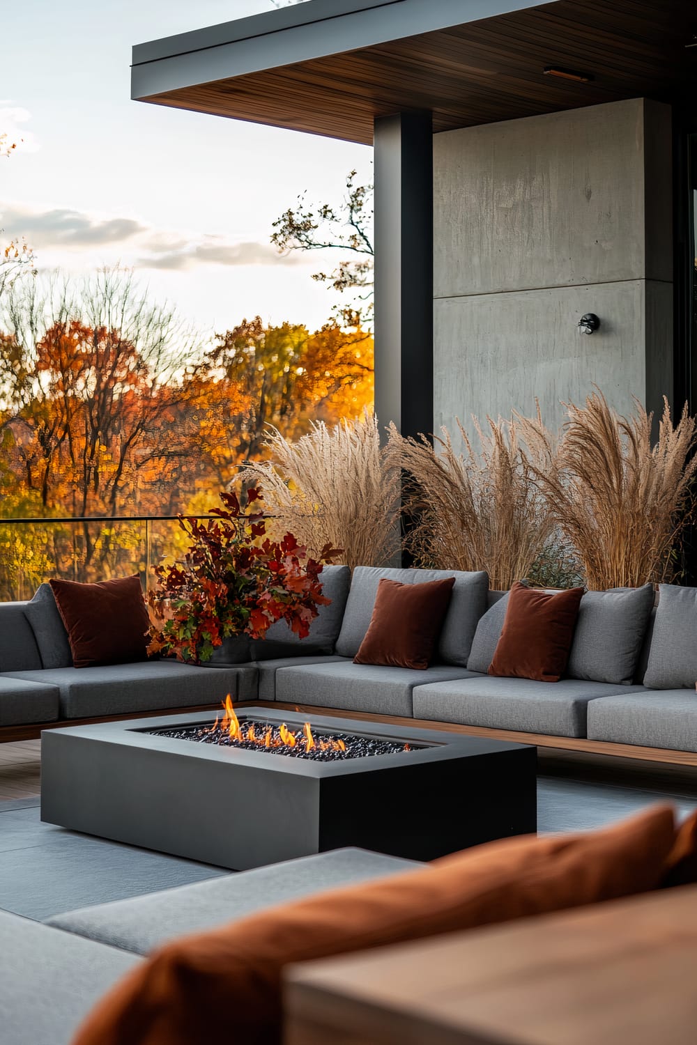 A modern outdoor patio featuring a sectional gray sofa with rust-colored throw pillows arranged around a sleek black fire pit. Ornamental grasses in planters add texture to the setting. The background showcases fall foliage with trees displaying vibrant autumn colors such as orange, yellow, and red.