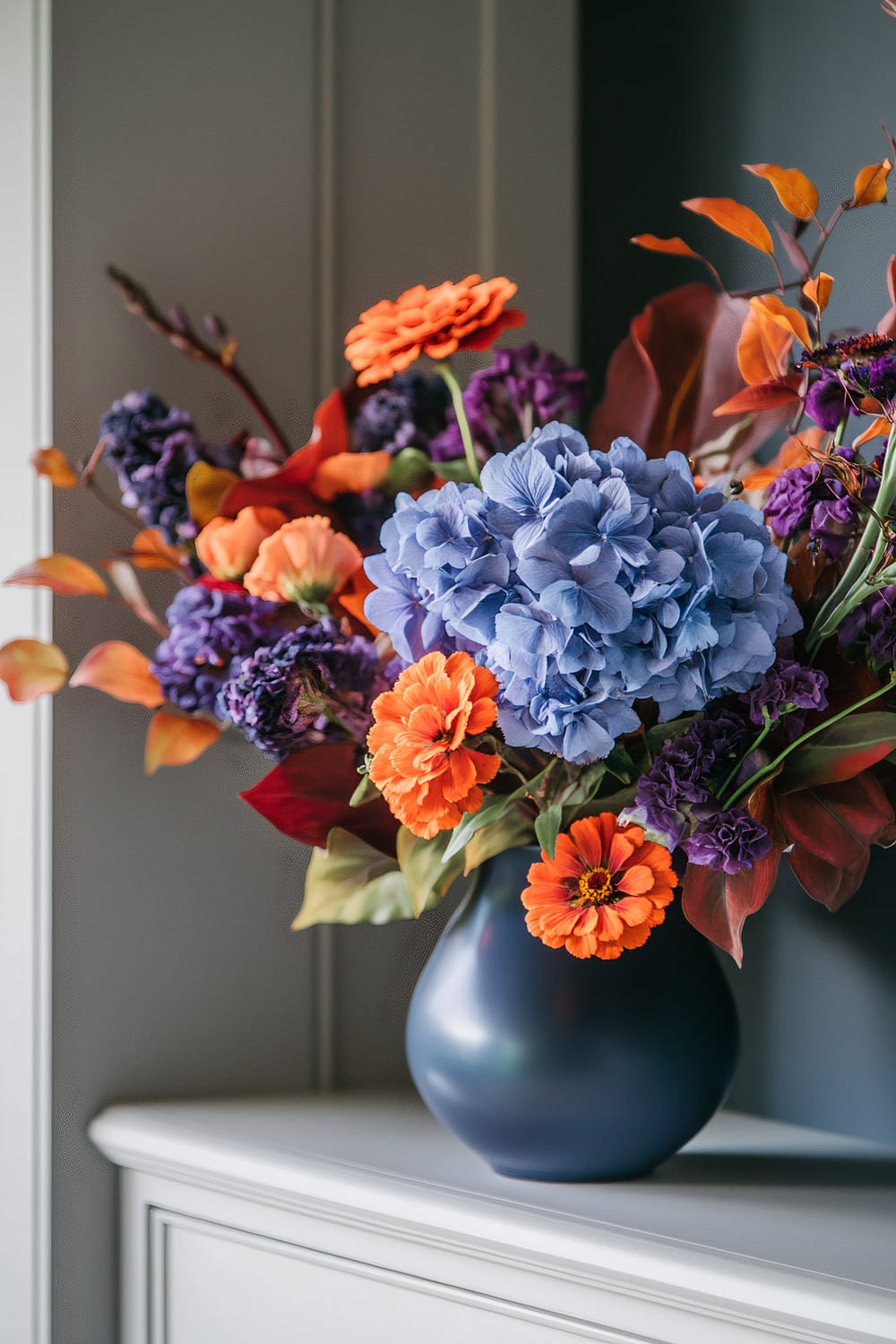 A striking bouquet of vibrant flowers arranged in a deep blue vase is displayed on a shelf. The bouquet features a mix of orange zinnias, blue hydrangeas, and deep purple flowers, with autumnal leaves adding a touch of warm tones. It is set against a neutral gray background.