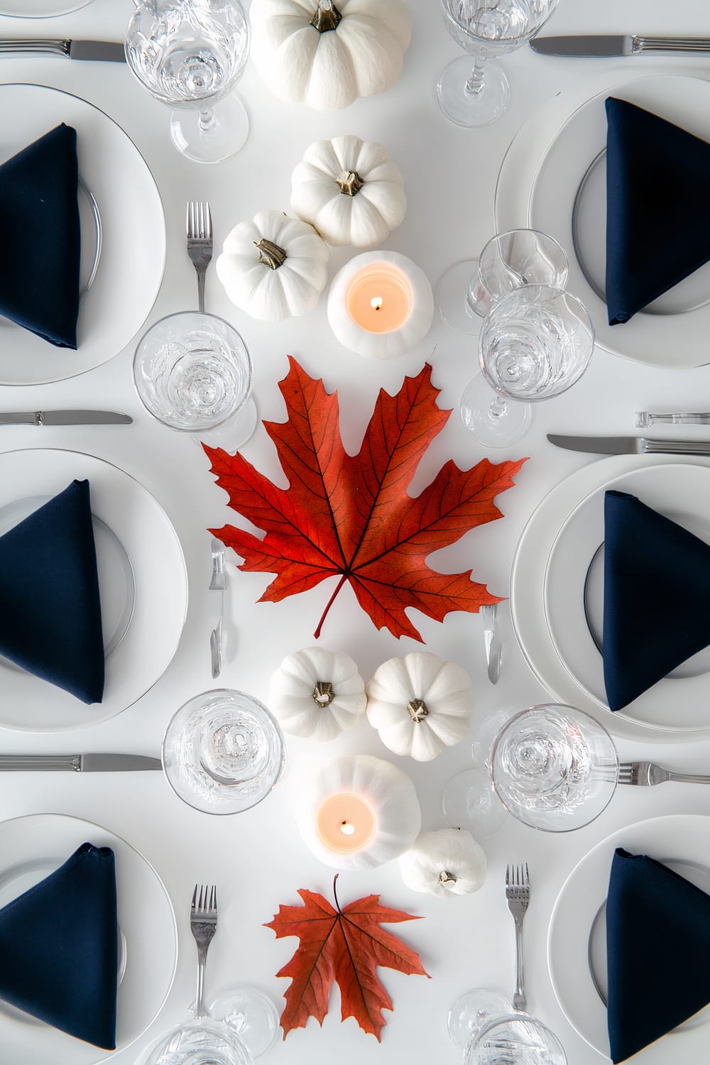 An elegant table setting for a fall-themed meal features white pumpkins, a central lit candle, and vivid red autumn leaves as the centerpiece. Each place setting includes white plates, silver cutlery, crystal glassware, and navy blue napkins neatly folded into triangles.