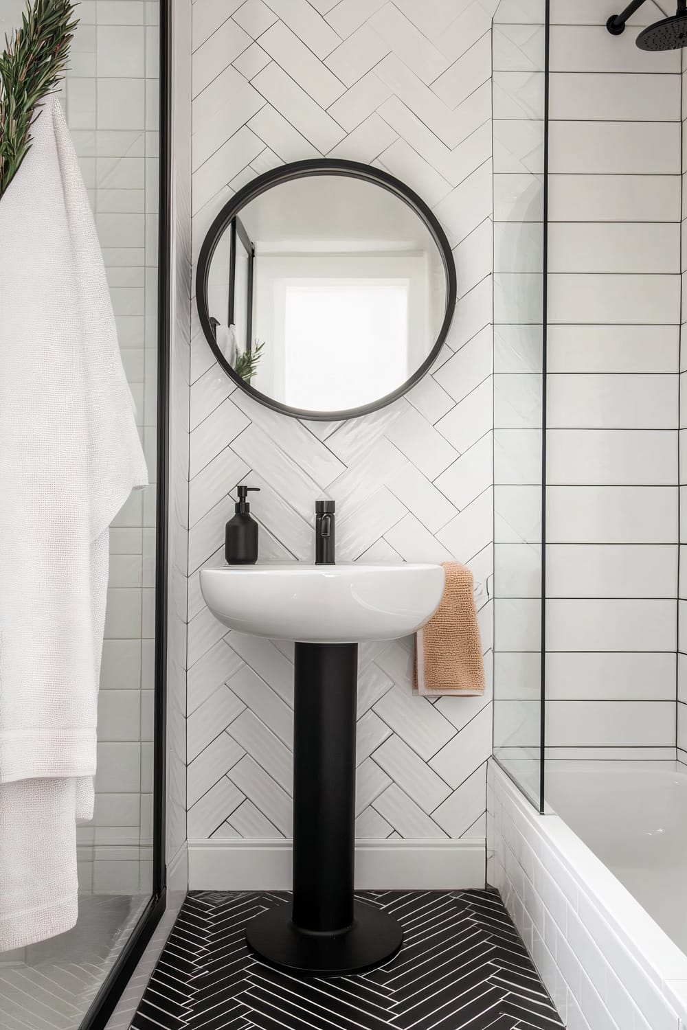 A modern bathroom featuring herringbone-patterned white wall tiles behind a sleek, round mirror with a black frame. Below the mirror, a minimalist pedestal sink with a black base stands on a visually striking black herringbone-tiled floor. To the left, a white towel hangs from a black-framed glass shower partition, while a small, beige towel is draped on a silver towel holder to the right. The shower area and bathtub are enclosed with glass panels, maintaining an airy and open feel.